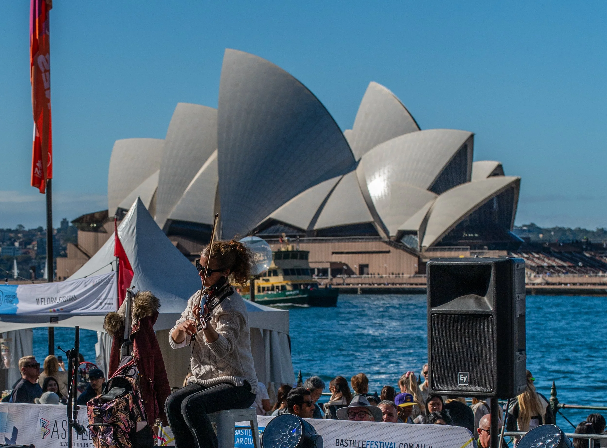 A musician playing the violin outdoors near the Sydney Opera House on a sunny day with a crowd of people watching, and boats on the water in the background.