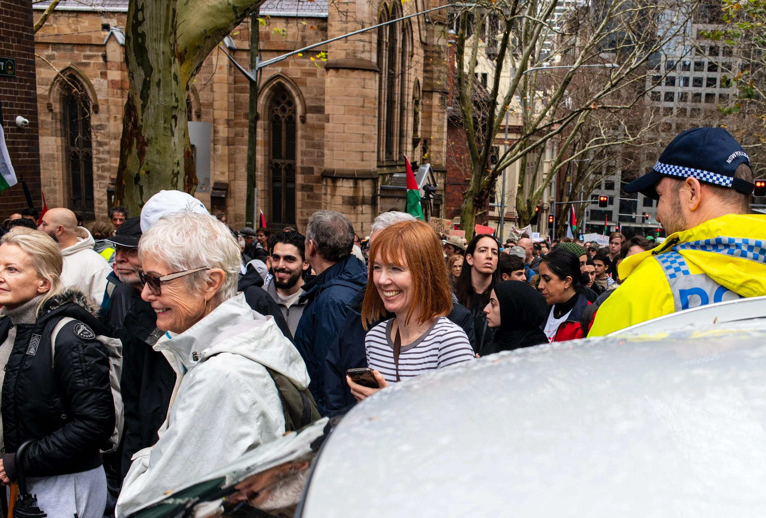 A diverse group of people participating in a protest or rally, with some holding signs and flags, in an outdoor urban setting with trees and historic buildings.