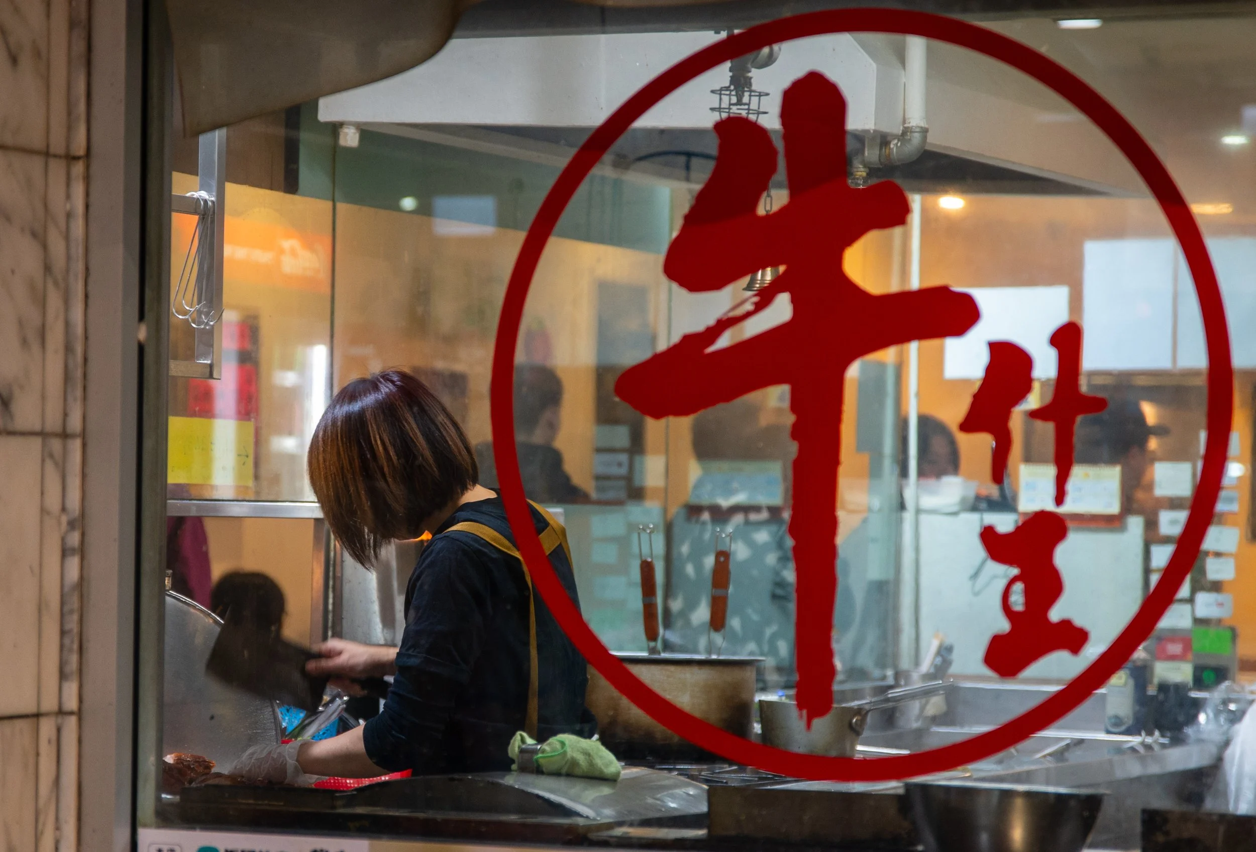 A woman preparing food in a restaurant kitchen viewed through a glass window with a red Chinese character sign.