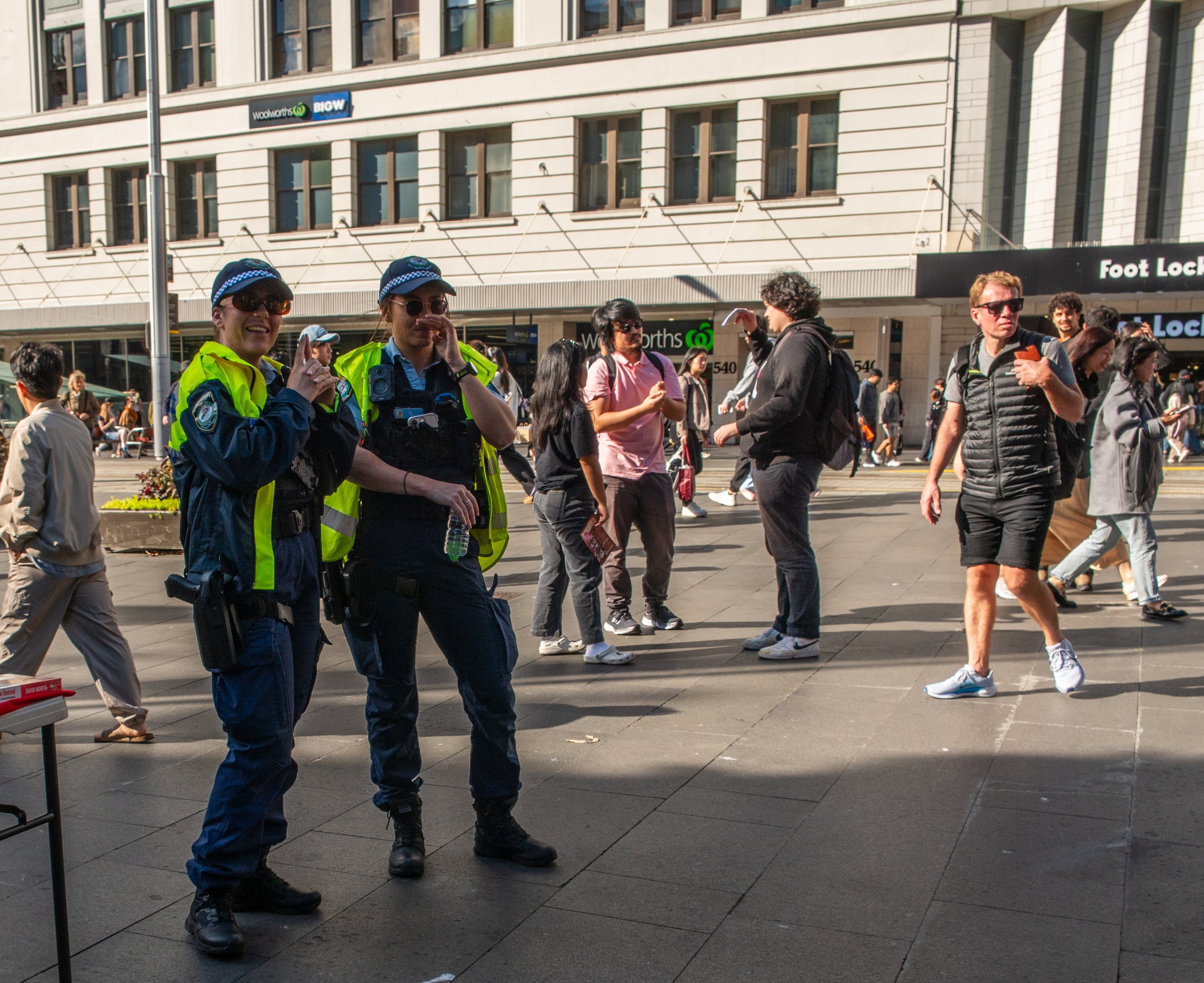 Two police officers in uniform and reflective vests standing on a busy city sidewalk, with pedestrians walking in various directions in the background.
