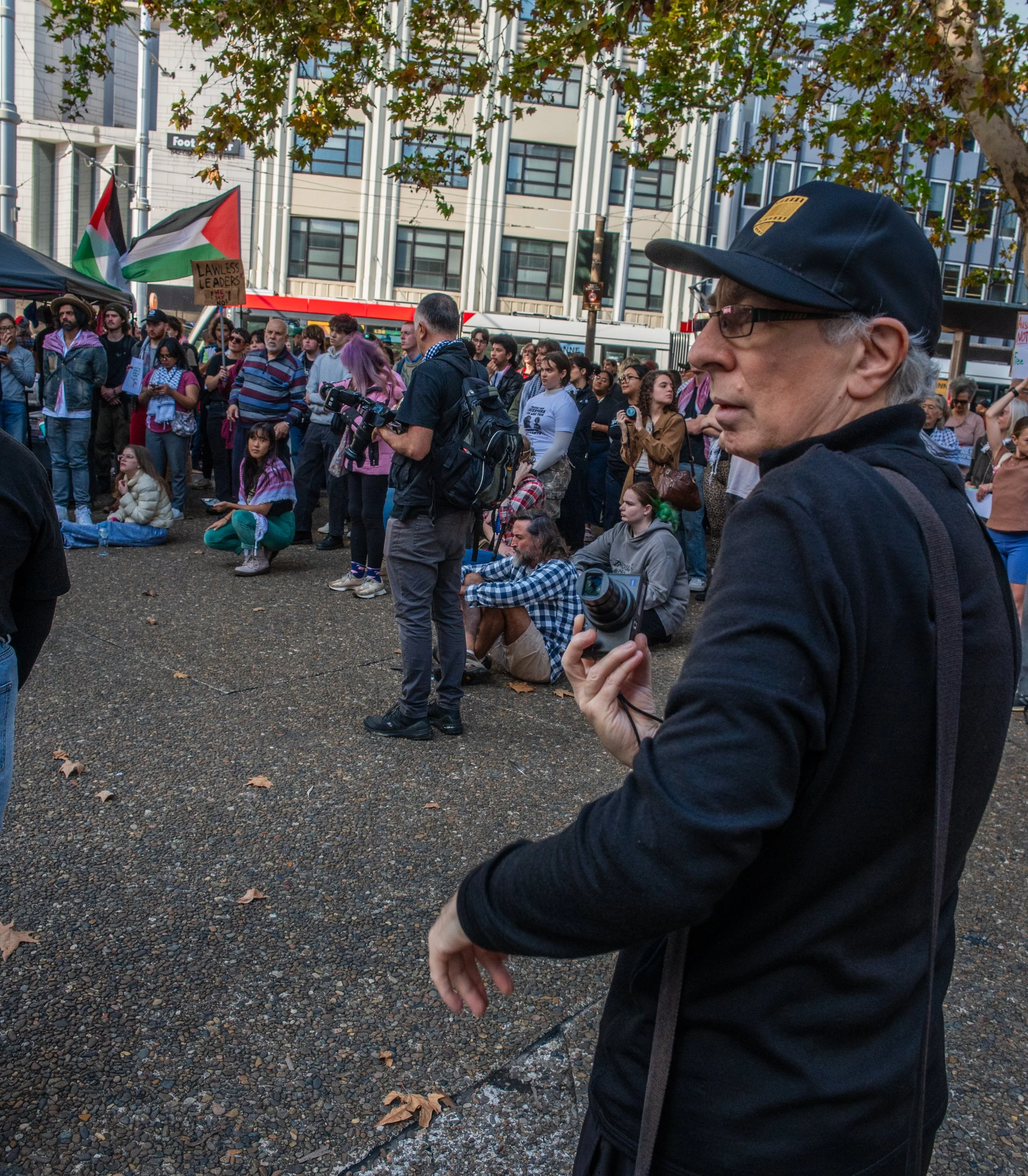 Crowd of protesters gathered outdoors, some sitting and some standing, with flags including a Palestinian flag, in front of modern buildings. A man in the foreground holds a camera and wears a black hat and glasses.