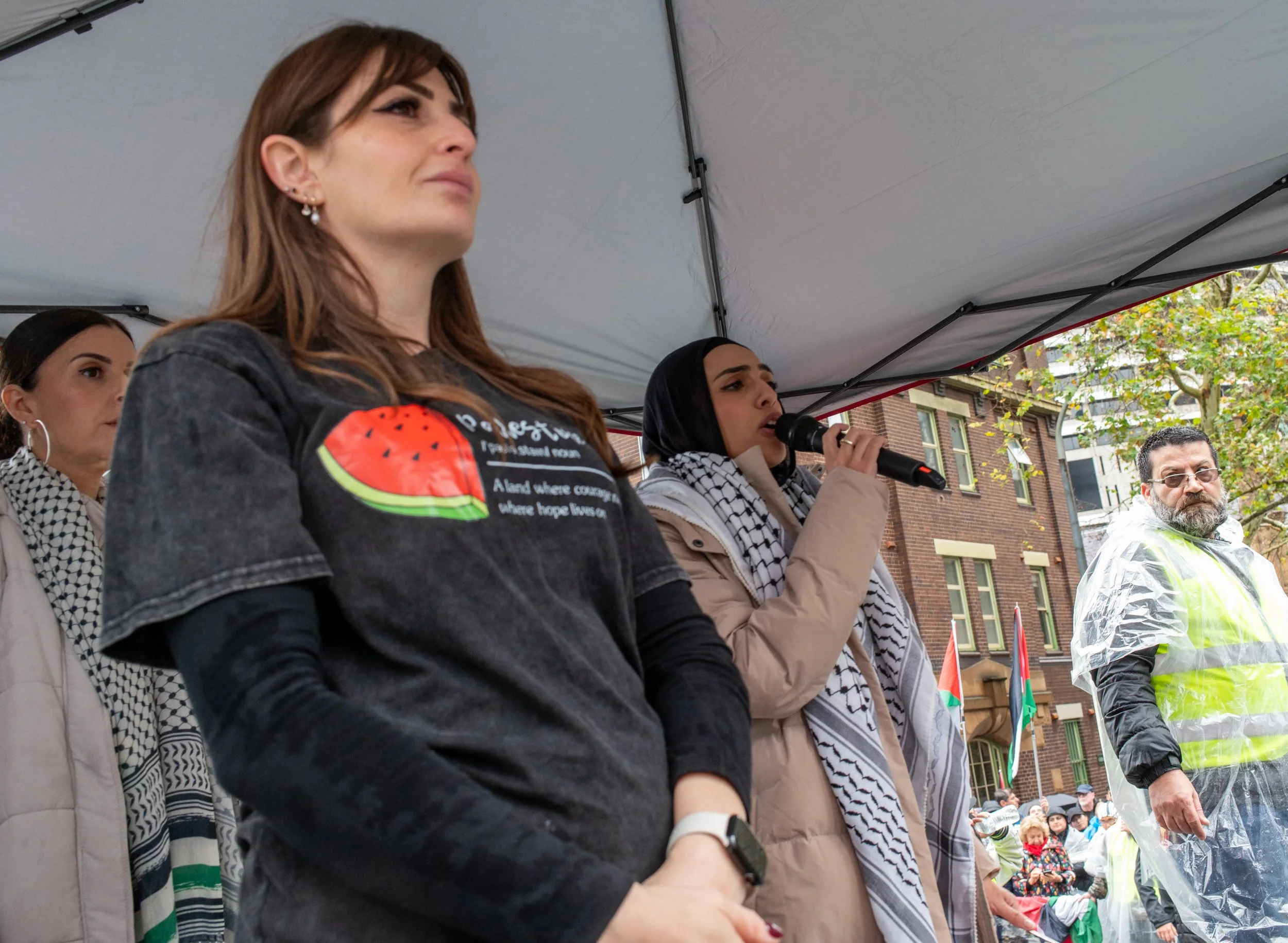 Women speaking at a protest or rally under a tent, with people holding flags and wearing rain gear in the background.