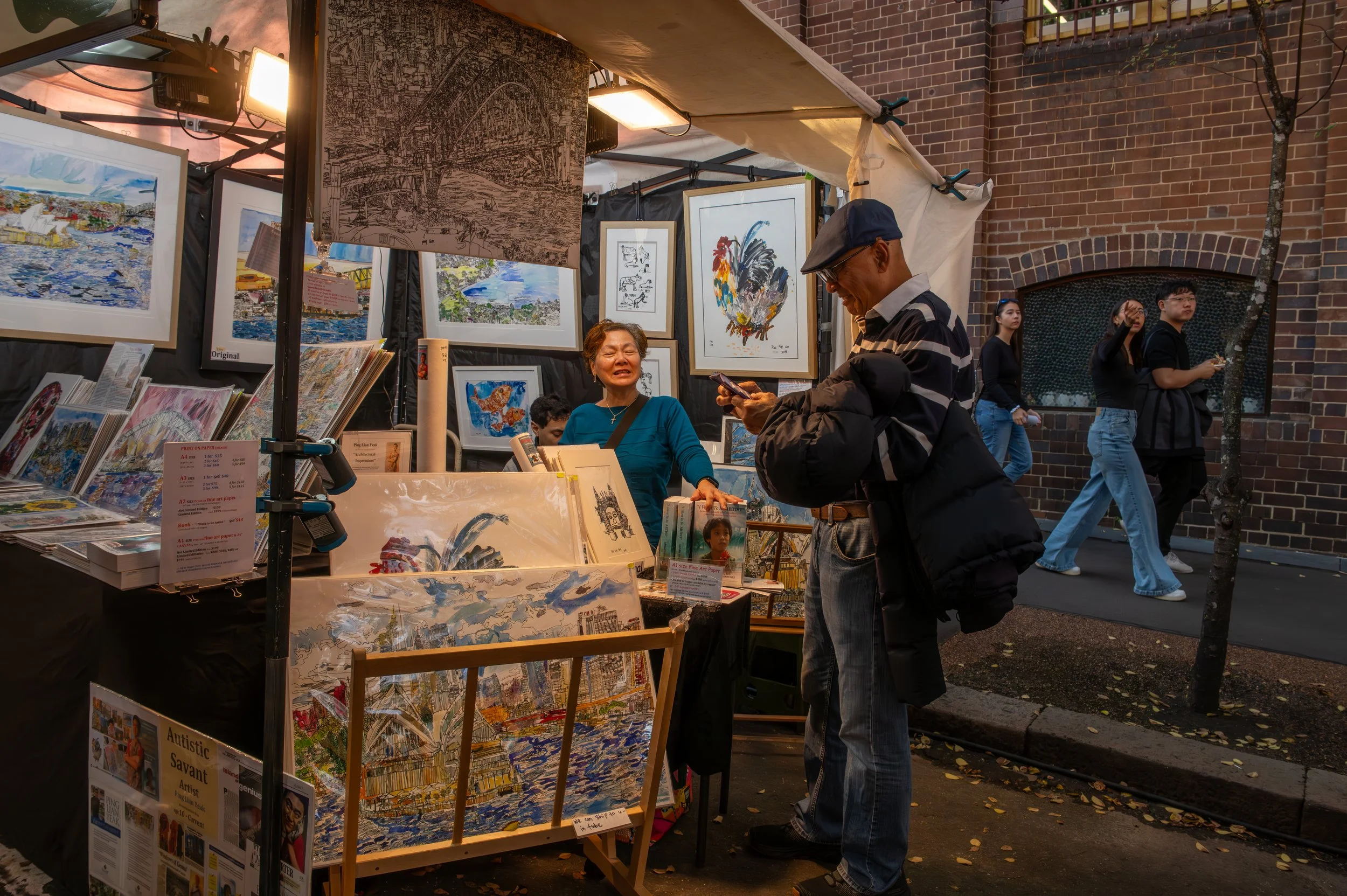 An outdoor art market at dusk with paintings on display, a woman selling art, and a man looking at his phone, with other pedestrians walking by.