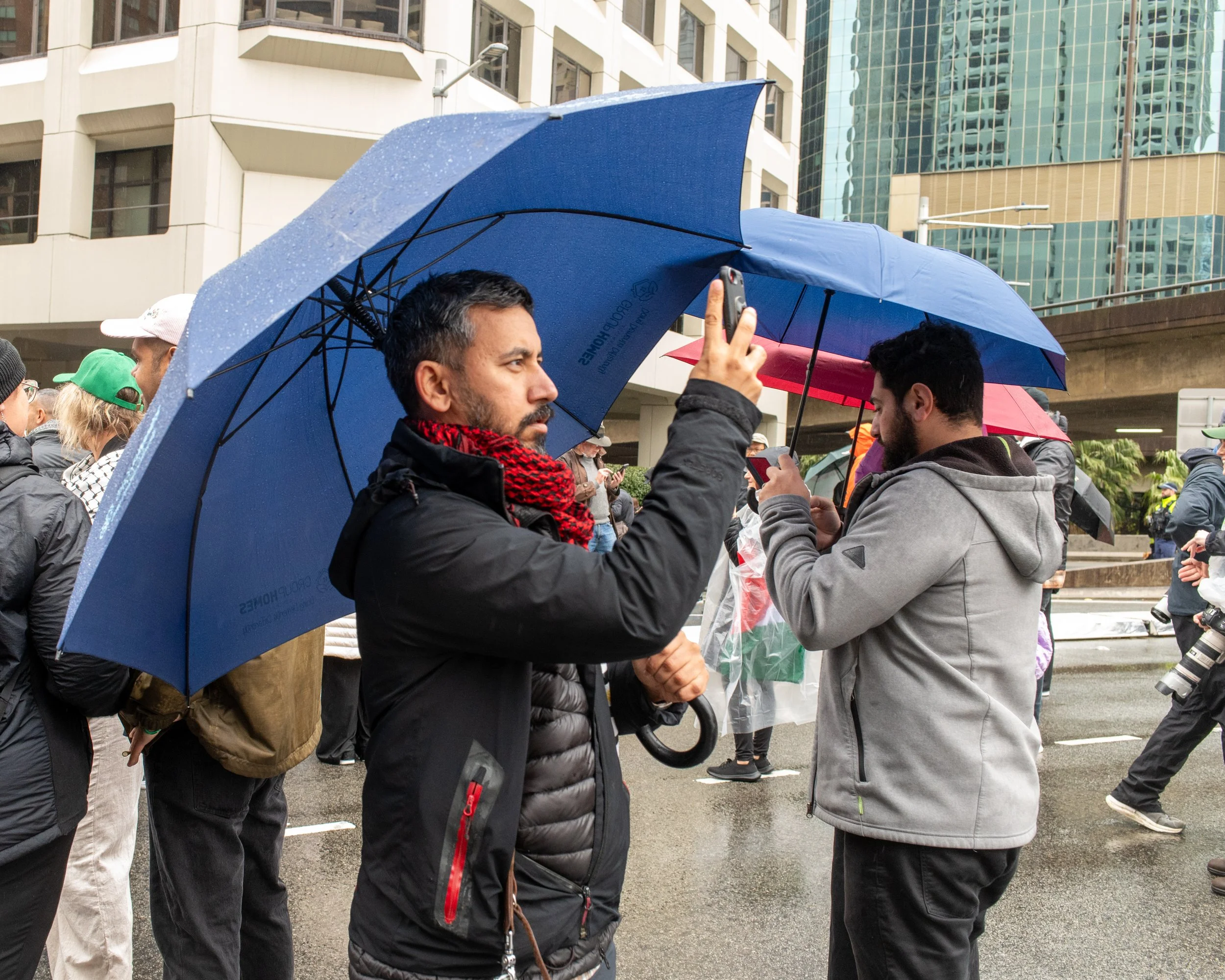 Two men under umbrellas taking photos or videos with their smartphones during a rainy day protest or gathering in an urban setting.