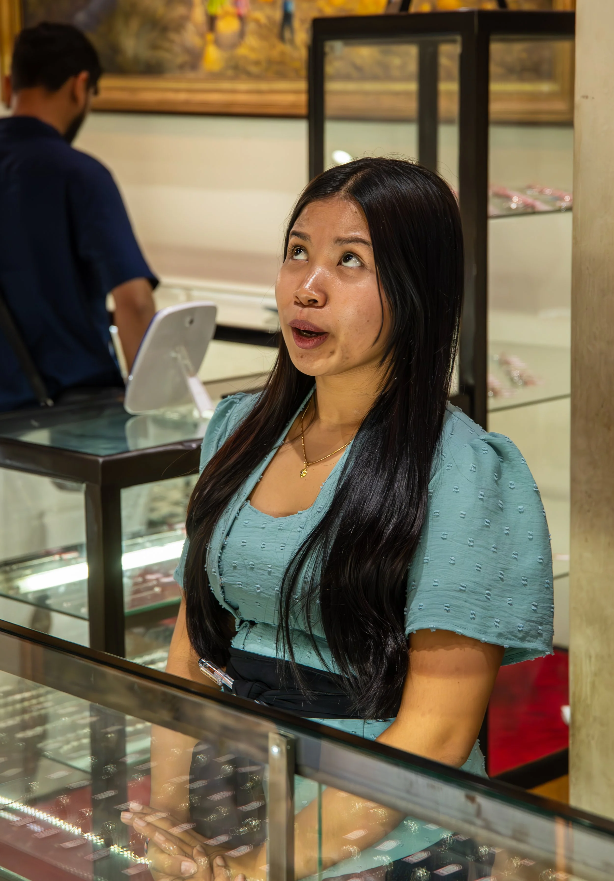 A woman with long black hair wearing a light blue blouse, standing inside a jewelry store, looking up with a surprised or curious expression. In the background, a man stands behind a glass display case filled with jewelry.