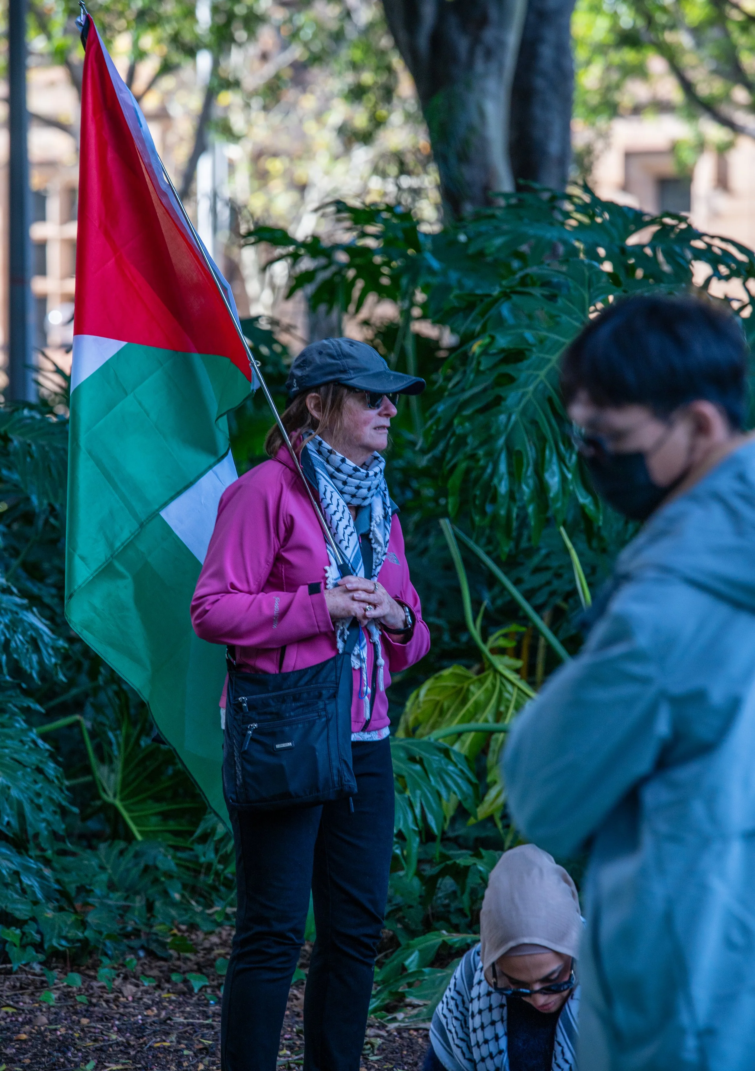 A woman holding a Palestinian flag standing outdoors next to a woman in a beige headscarf and another man wearing a black face mask, in front of green leafy plants.