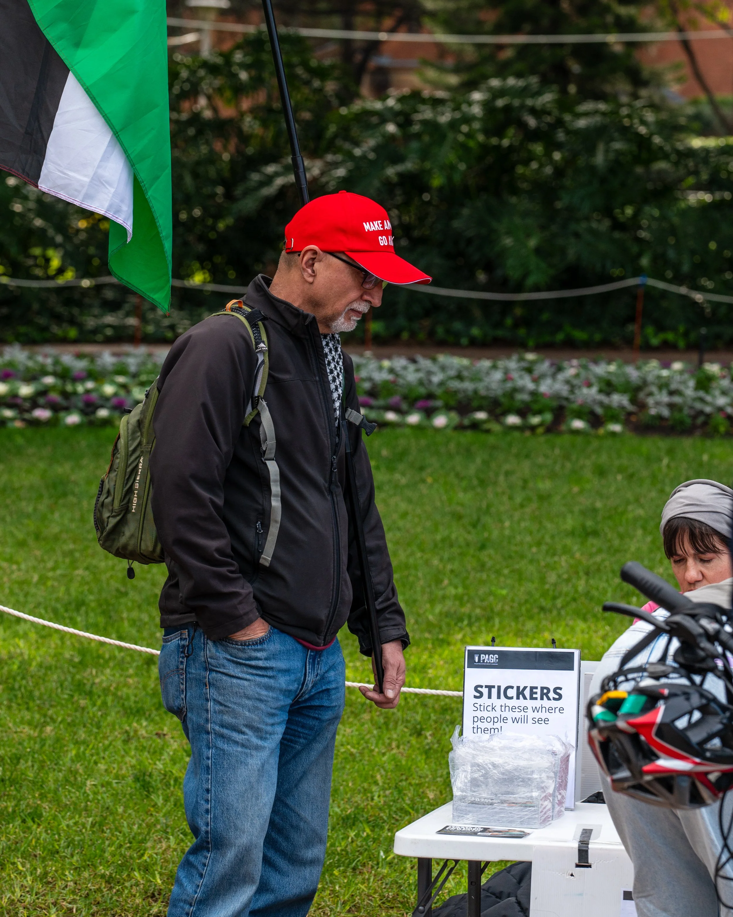 Man wearing a red Make America Great Again hat, black jacket, blue jeans, and backpack, looking at stickers on a table labeled 'STICKERS' with instructions to stick them where people will see them, outdoors on grass with flowers and trees in the background.
