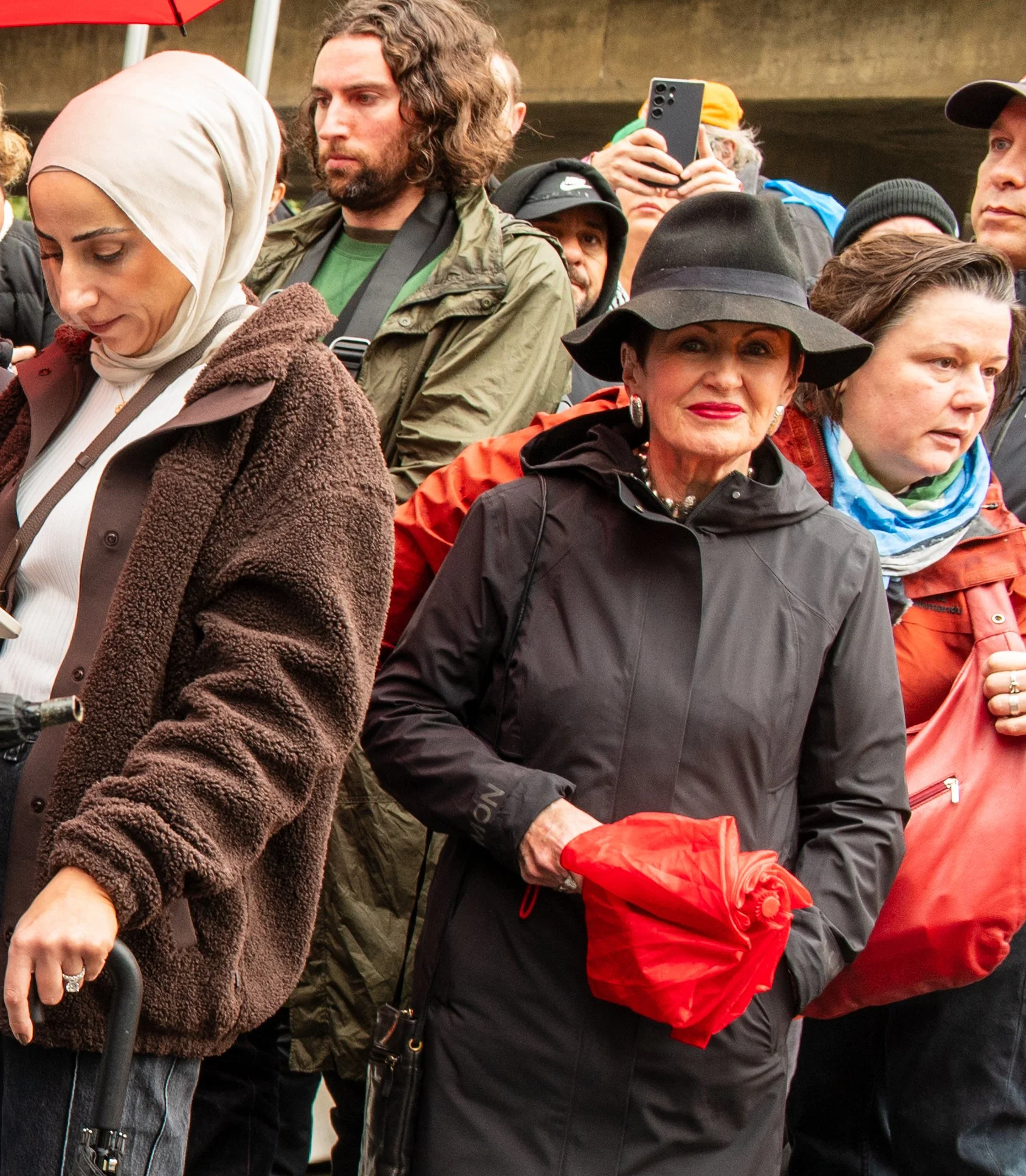 A group of people gathered outdoors, some holding umbrellas and wearing jackets. Prominent in the center is an older woman wearing a wide-brimmed black hat, a black jacket, and holding a red bag.