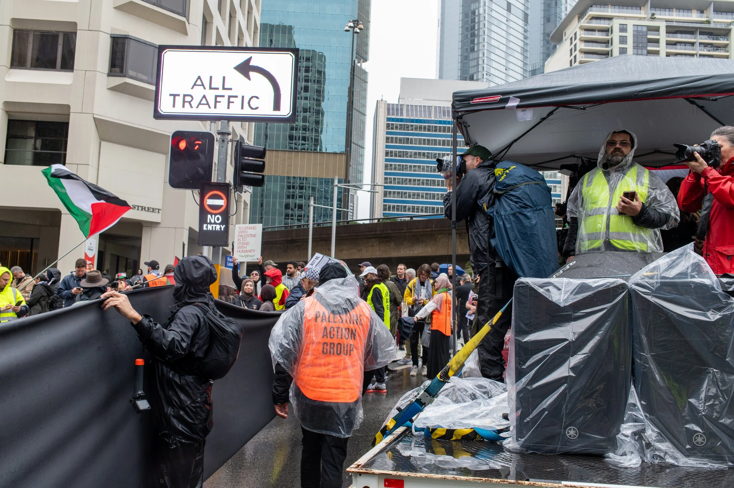 A crowd gathers at a protest on a city street, with people holding flags and signs. Some individuals are wearing rain ponchos and high-visibility vests, and there is a press presence with cameras capturing the event. The scene appears rainy with wet pavement and people dressed for rain.