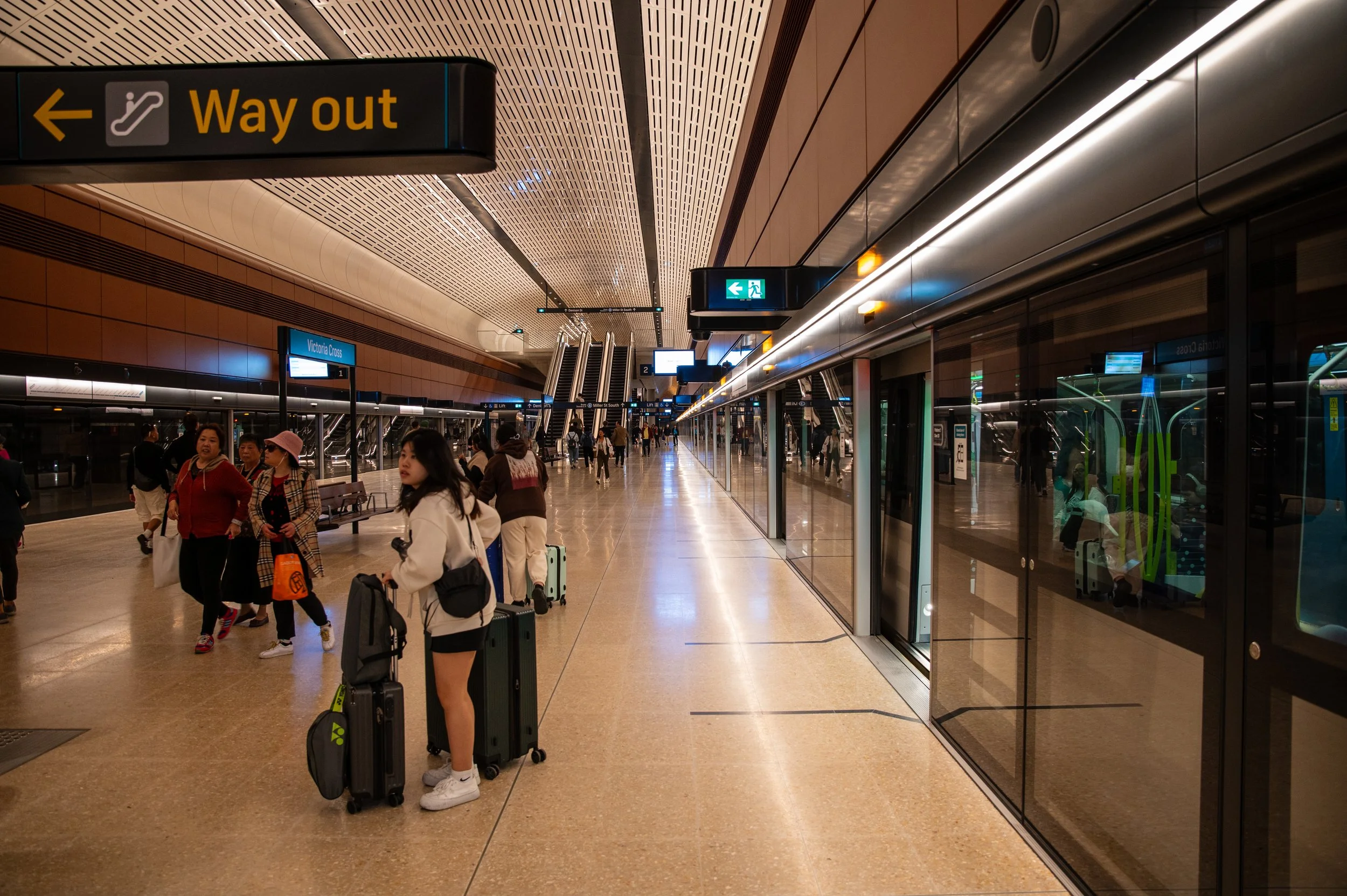 People with suitcases waiting in an airport terminal near a glass wall with yellow signs overhead directing to the way out and escalators in the background.