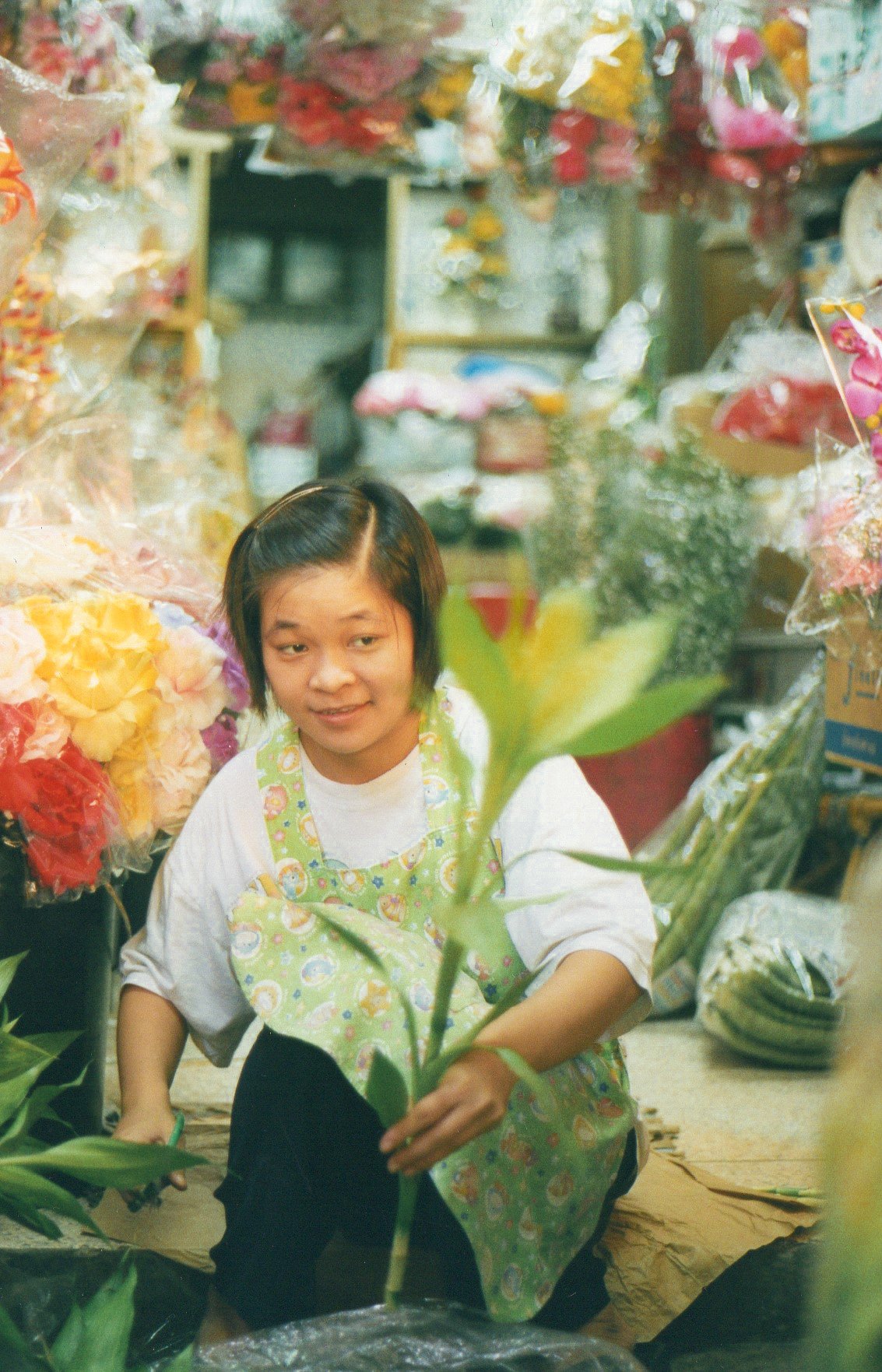 A woman working in a flower shop surrounded by colorful flowers and plants, holding a flower stem with leaves.