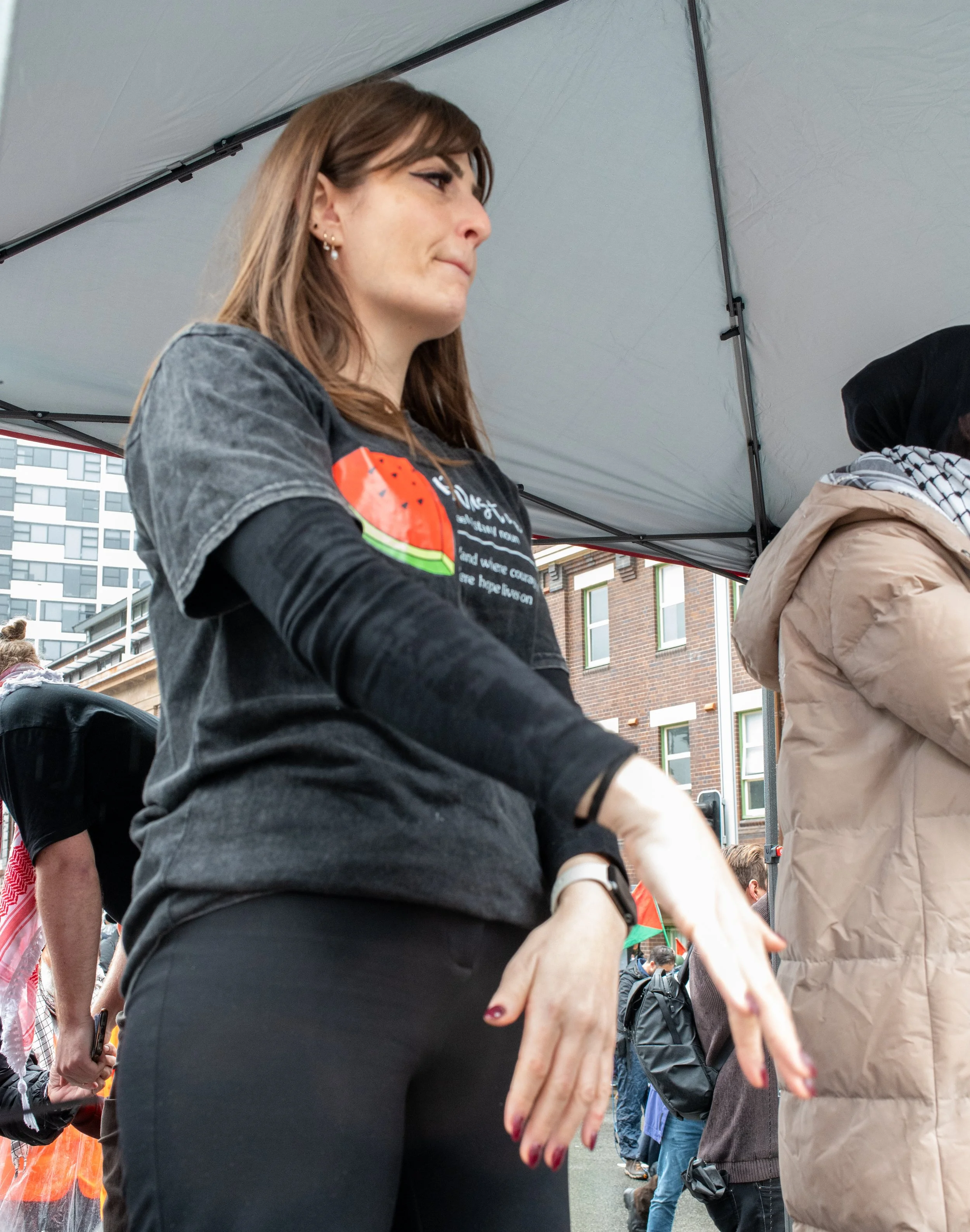 A woman with shoulder-length brown hair and earrings, wearing a black T-shirt and long-sleeve black top underneath, standing under a canopy at an outdoor event, with multiple people and buildings visible in the background.