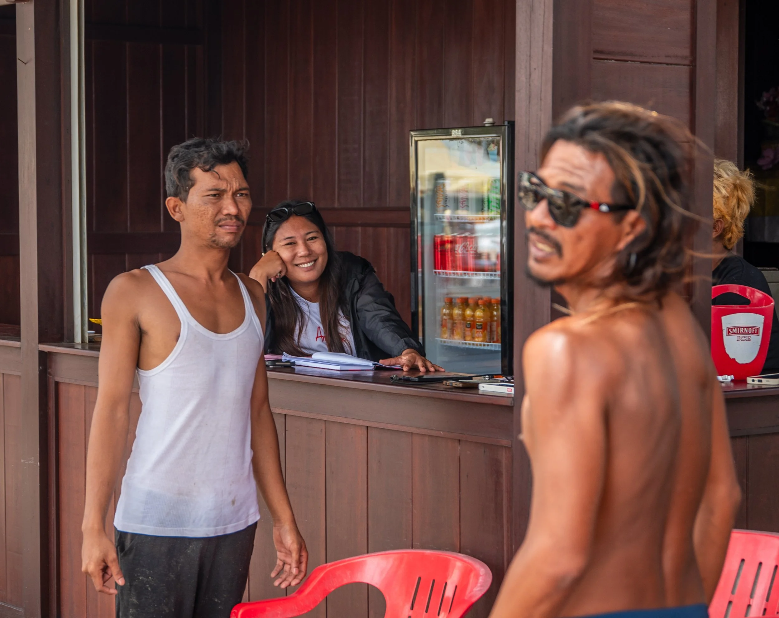 Three people are at an outdoor food stall, with two women sitting behind the counter and a woman in front talking to them. The woman behind the counter is smiling, and appears to be taking an order. The woman in the foreground is wearing sunglasses and no shirt, and appears to be talking or listening.