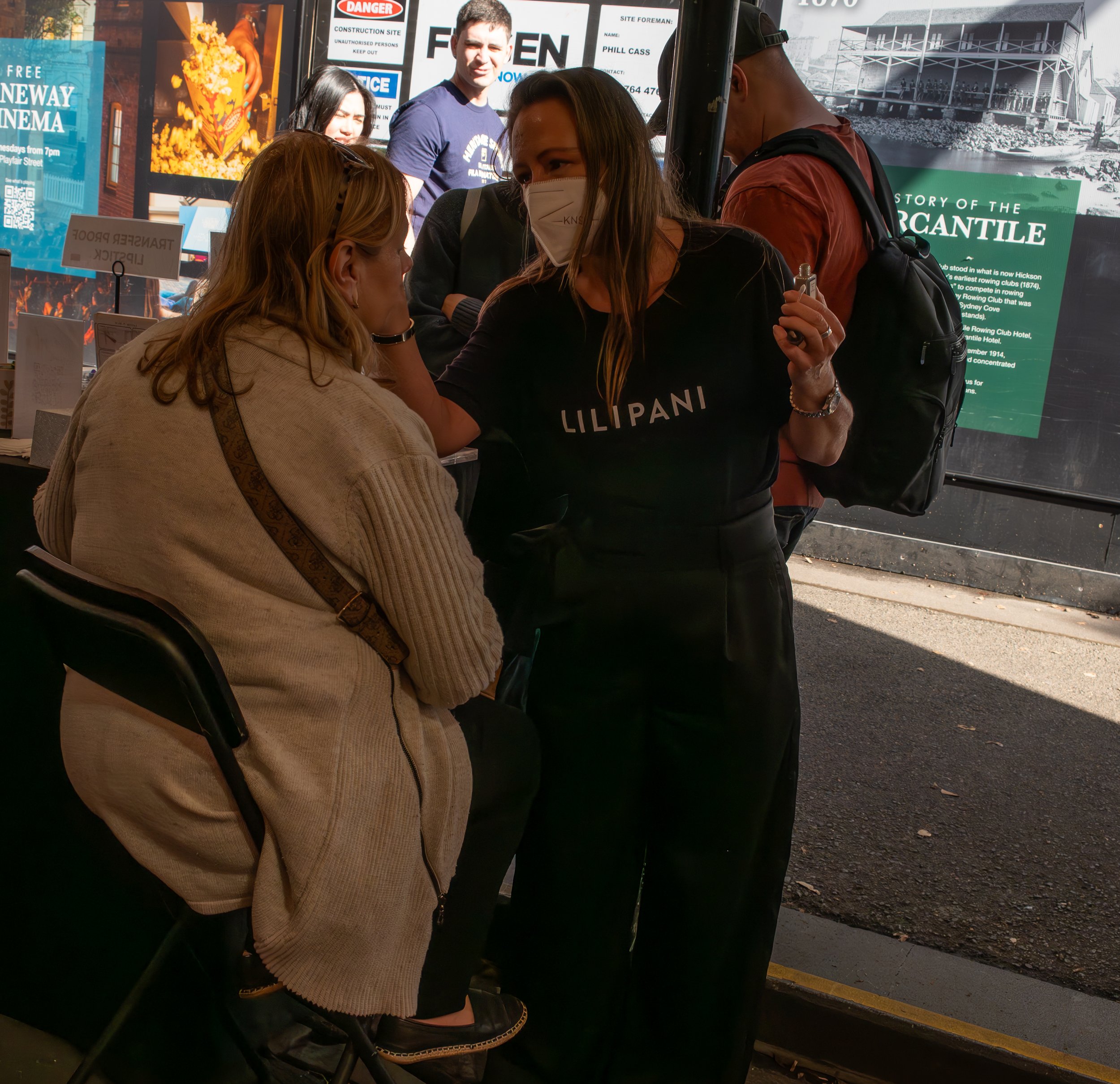 A woman wearing a black "LILLIPANI" t-shirt and a face mask applies makeup or face paint to a seated woman's face at an outdoor event or queue. Several people are visible in the background, with posters and signs on a wall behind them.