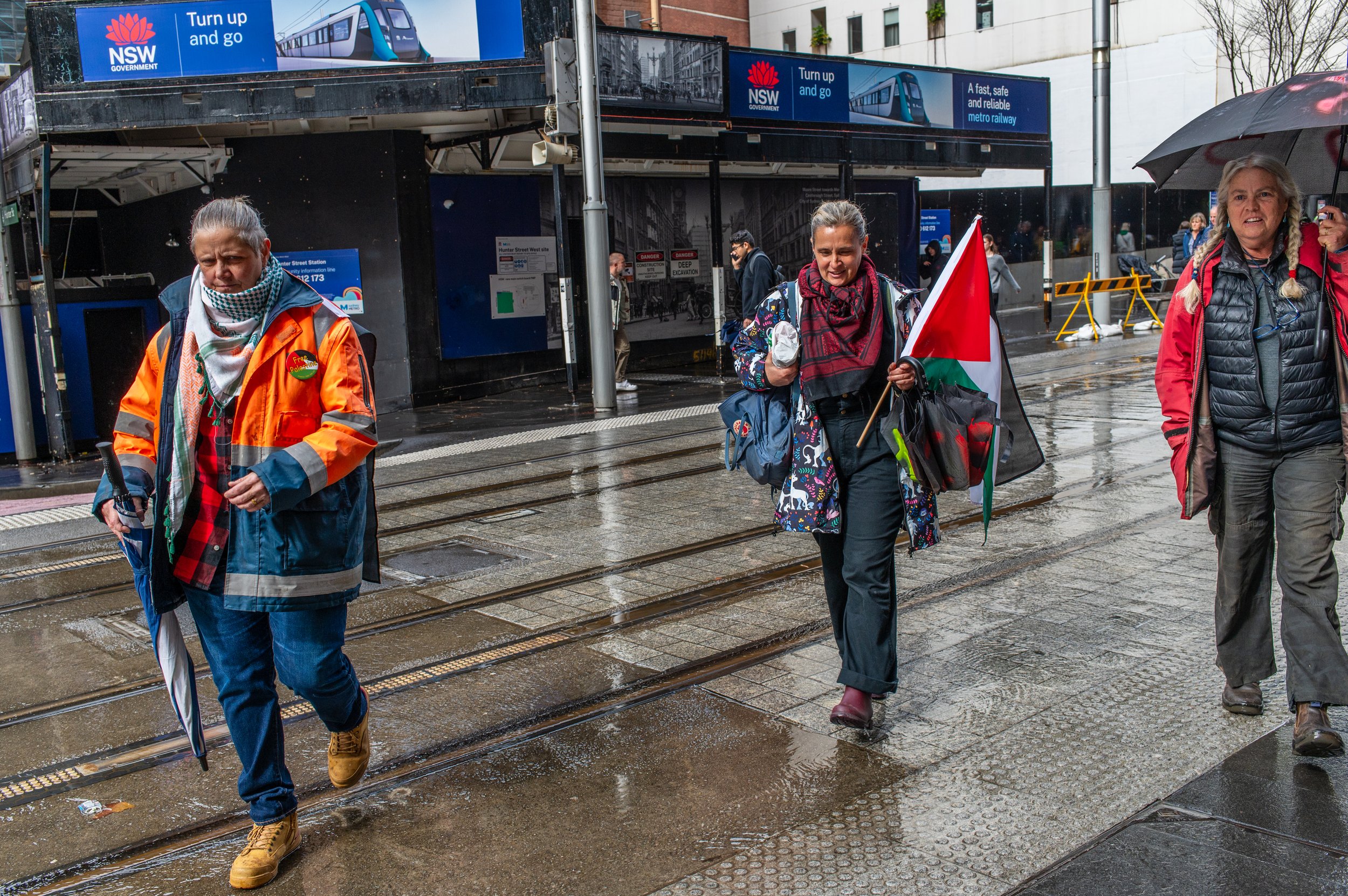 People walking on a wet city sidewalk, some holding umbrellas, with a subway station entrance in the background and political protests with flags.