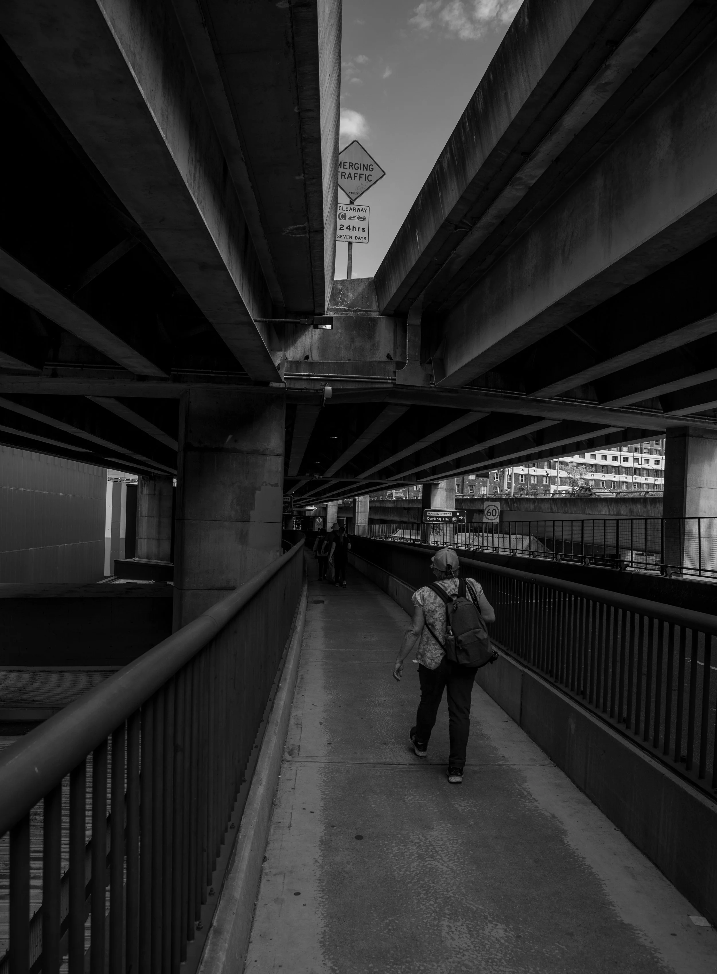Person walking on urban pedestrian walkway under overpasses, black and white photo.