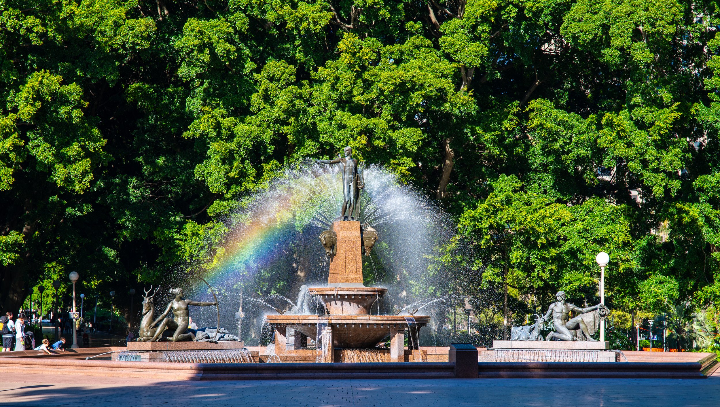 A fountain with sculptures of mythological figures, water spraying, and a rainbow against a backdrop of green trees in a park.
