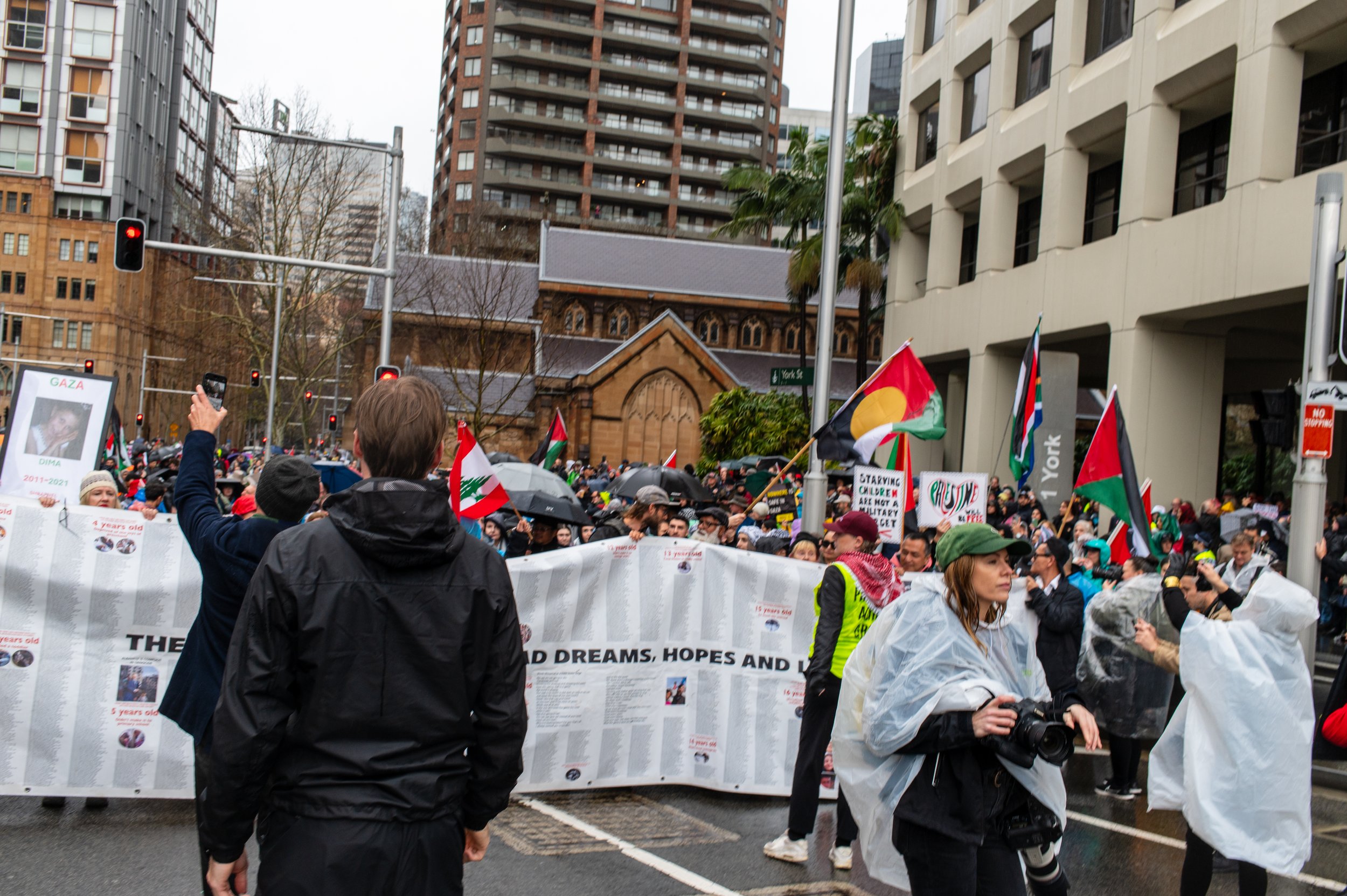 Crowd of protesters on a city street holding flags and banners, some wearing rain ponchos, with tall buildings and a church in the background.