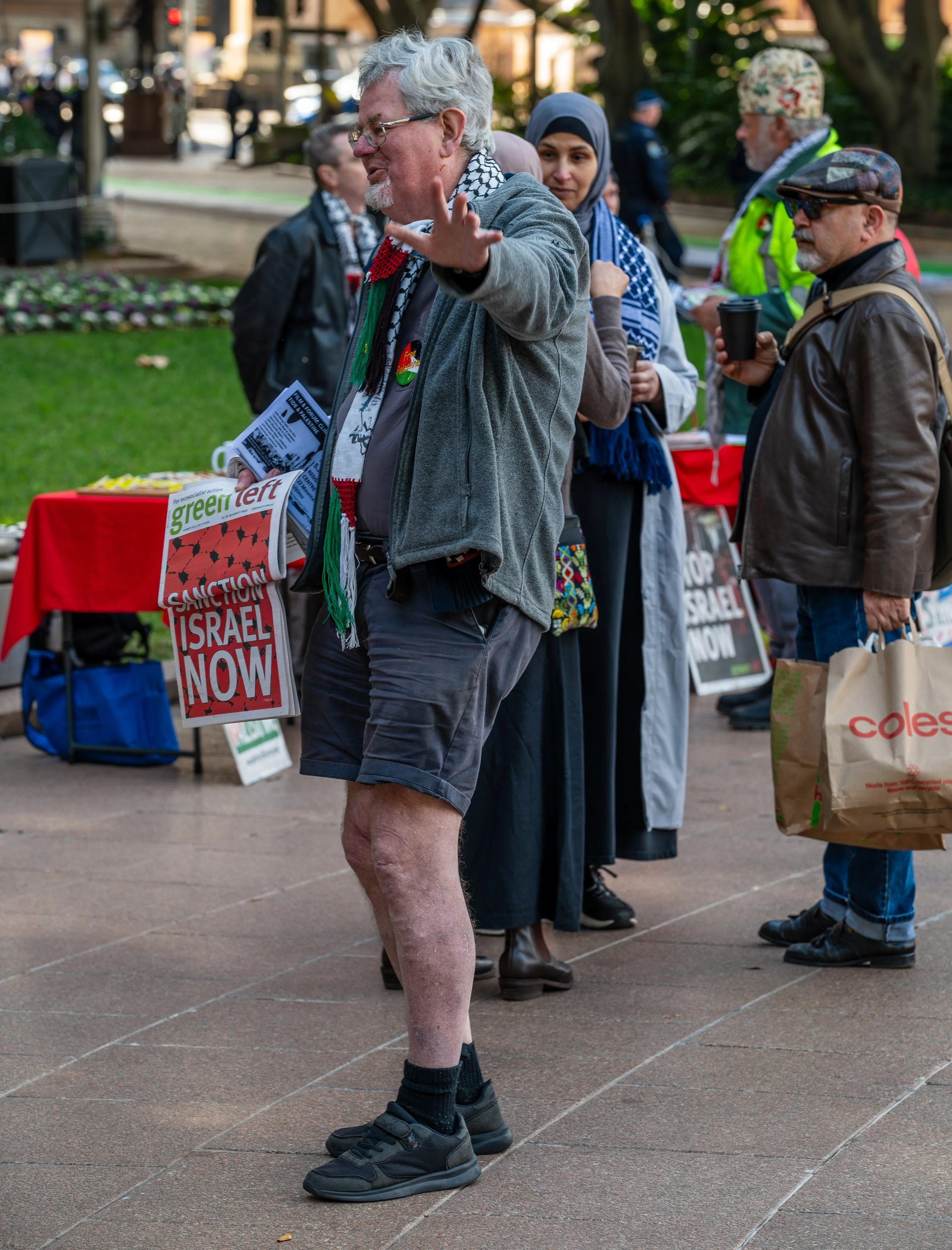 A man with gray hair, glasses, and a beard, wearing a gray jacket, shorts, and sneakers, is gesturing with his hand at a protest or rally. Behind him, several other people are holding signs and carrying bags, in a park or public space.
