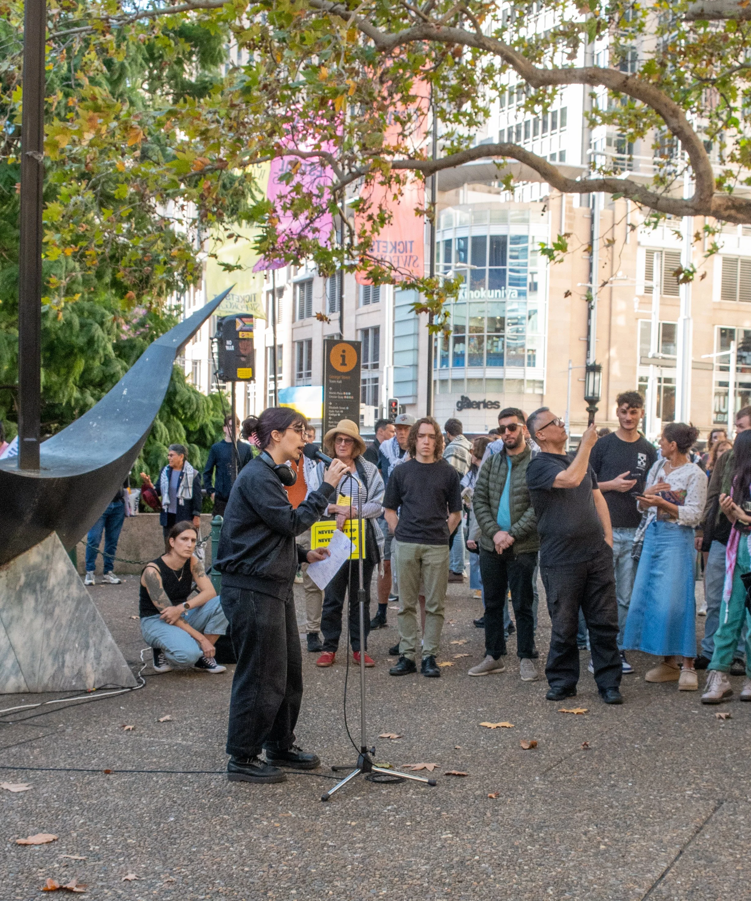 A woman speaking into a microphone at a public gathering in an urban park surrounded by a diverse group of people, some taking photos or listening attentively, with tall buildings and signs in the background.