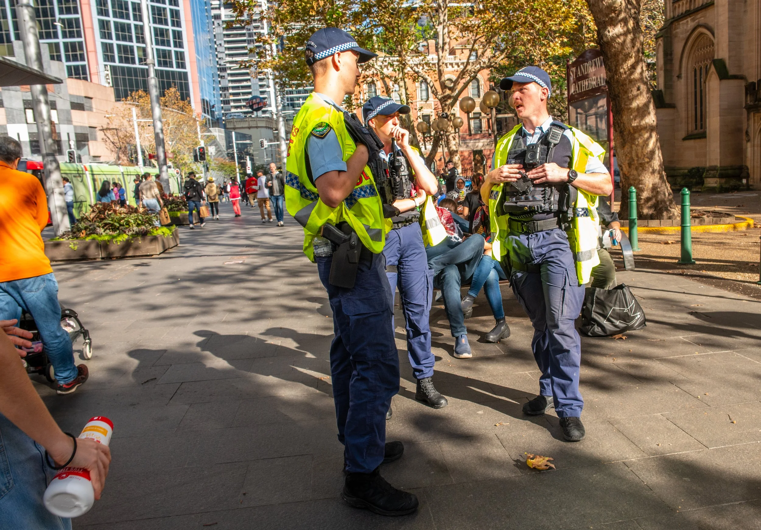 Three uniformed police officers wearing reflective vests are standing and talking on a city sidewalk, with pedestrians and buildings in the background.