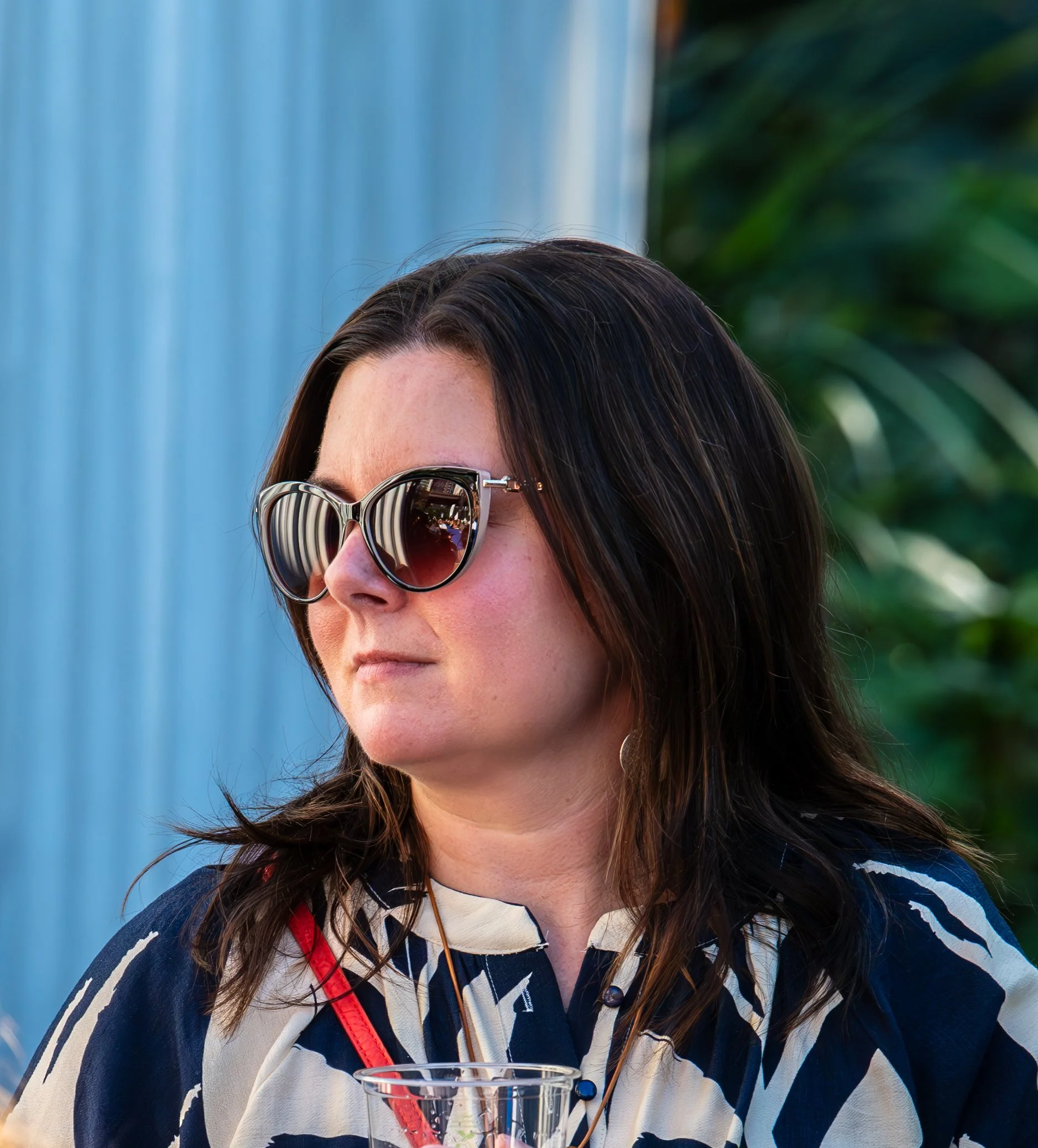 A woman with dark hair wearing sunglasses and a patterned shirt, holding a clear plastic cup, standing outdoors with green foliage and a blue fence in the background.