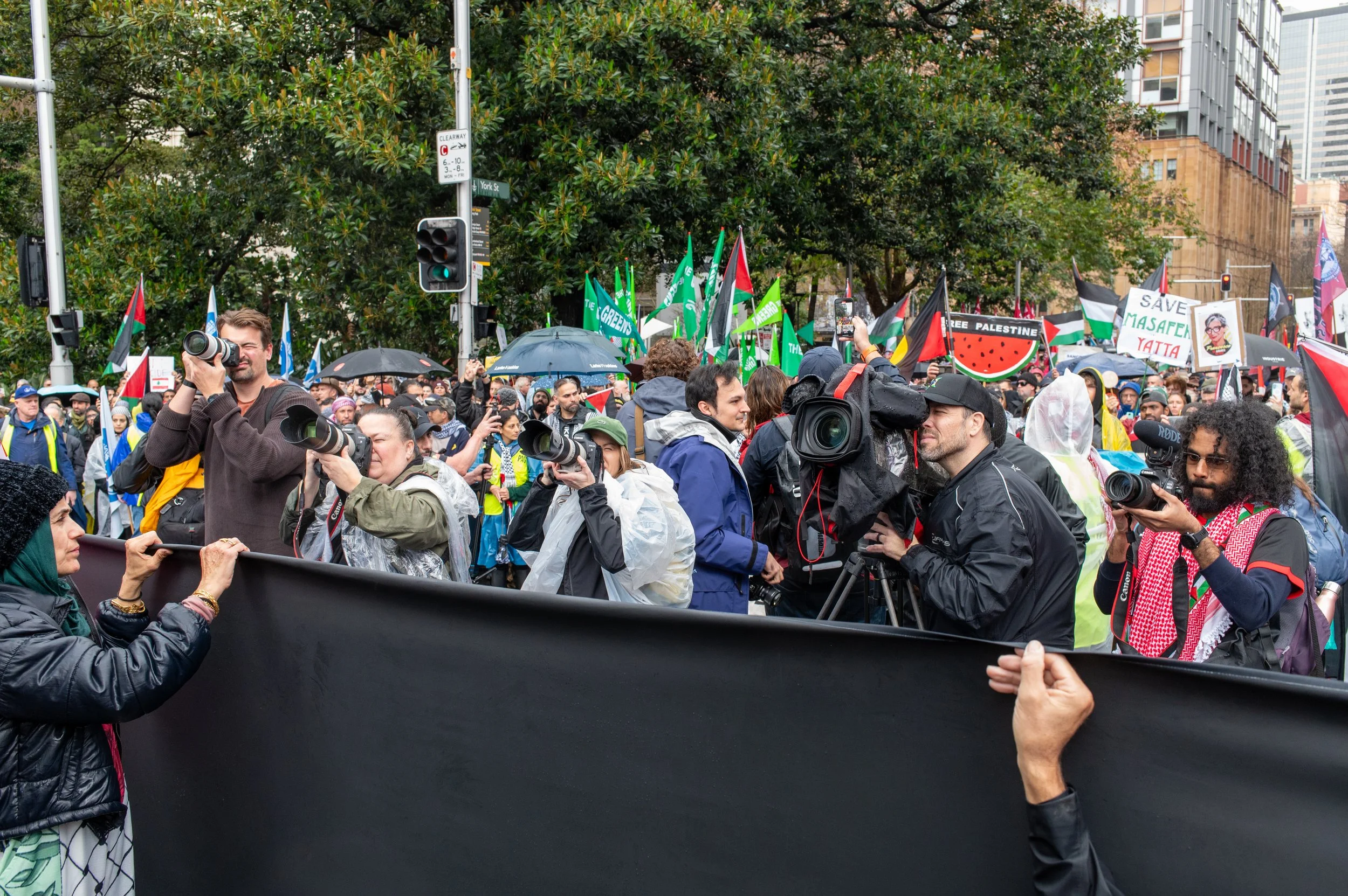 Crowd of protesters and journalists holding cameras at a demonstration, holding Palestinian flags and protest signs.