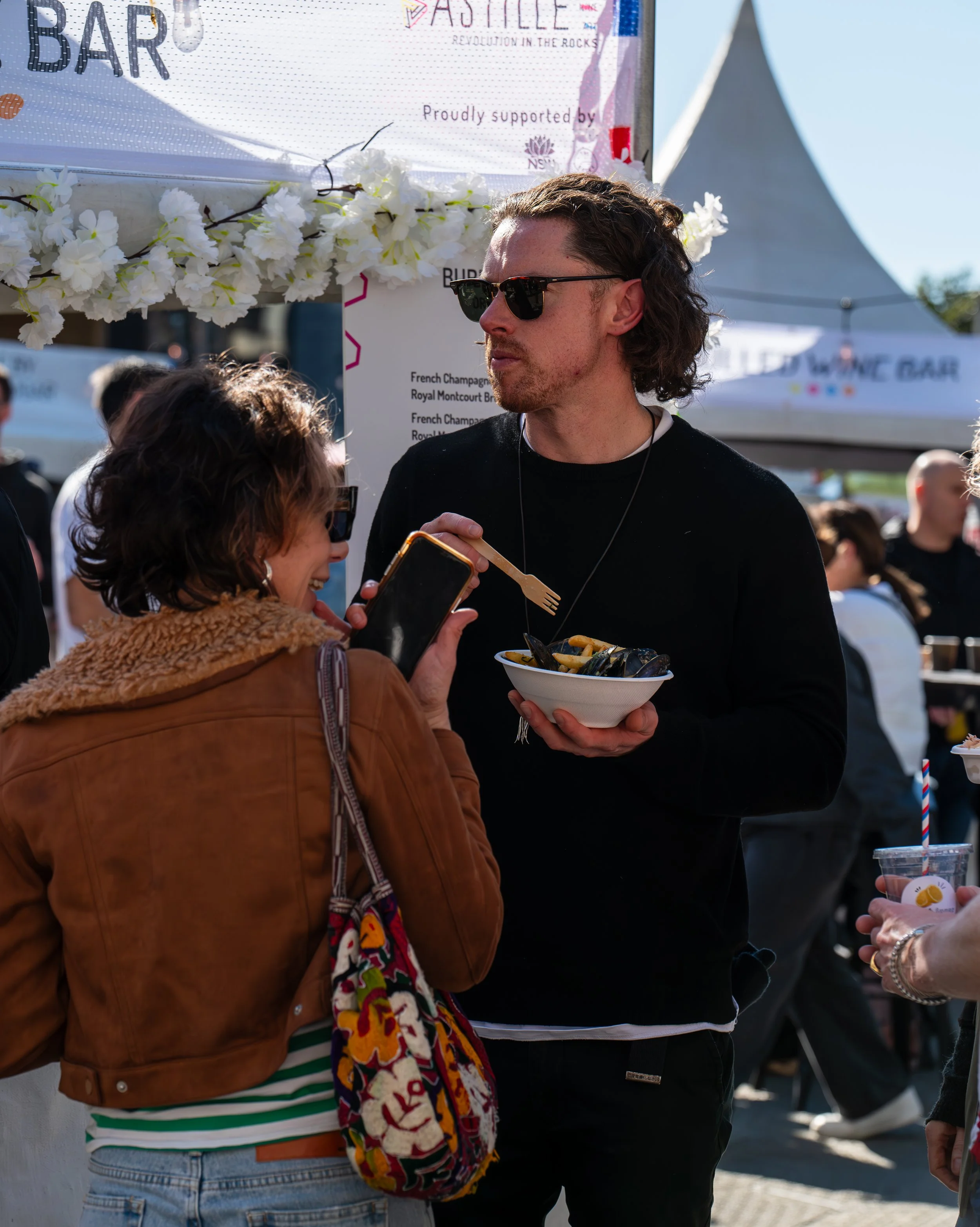 A man in black clothing with sunglasses holding a bowl of mussels during an outdoor event, talking to a woman with short brown hair, a brown jacket, and a colorful embroidered bag, taking a photo with her phone.