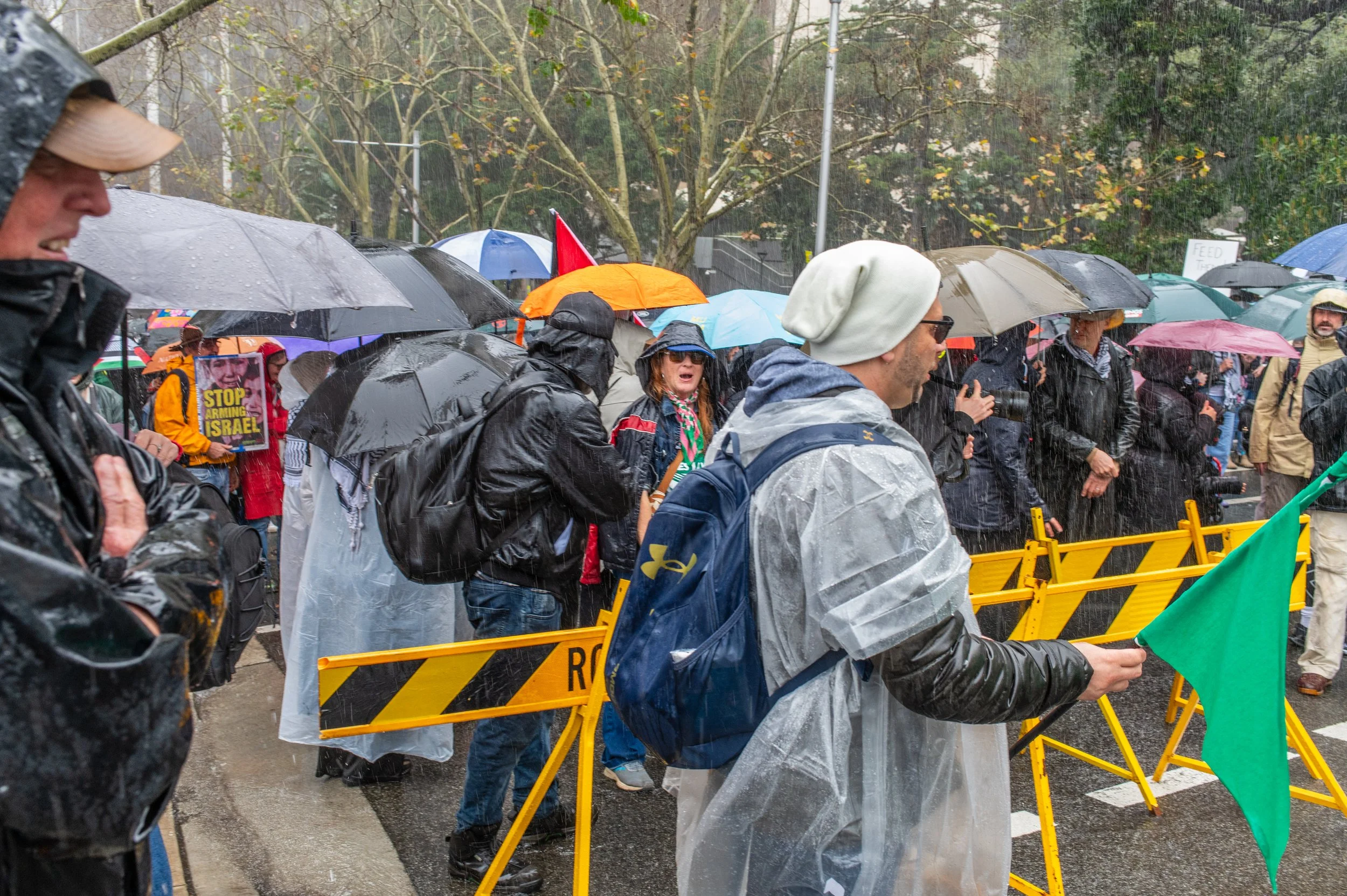 Crowd of people holding umbrellas, some wearing rain gear, protesting outdoors with yellow barricades and a green flag in rainy weather.