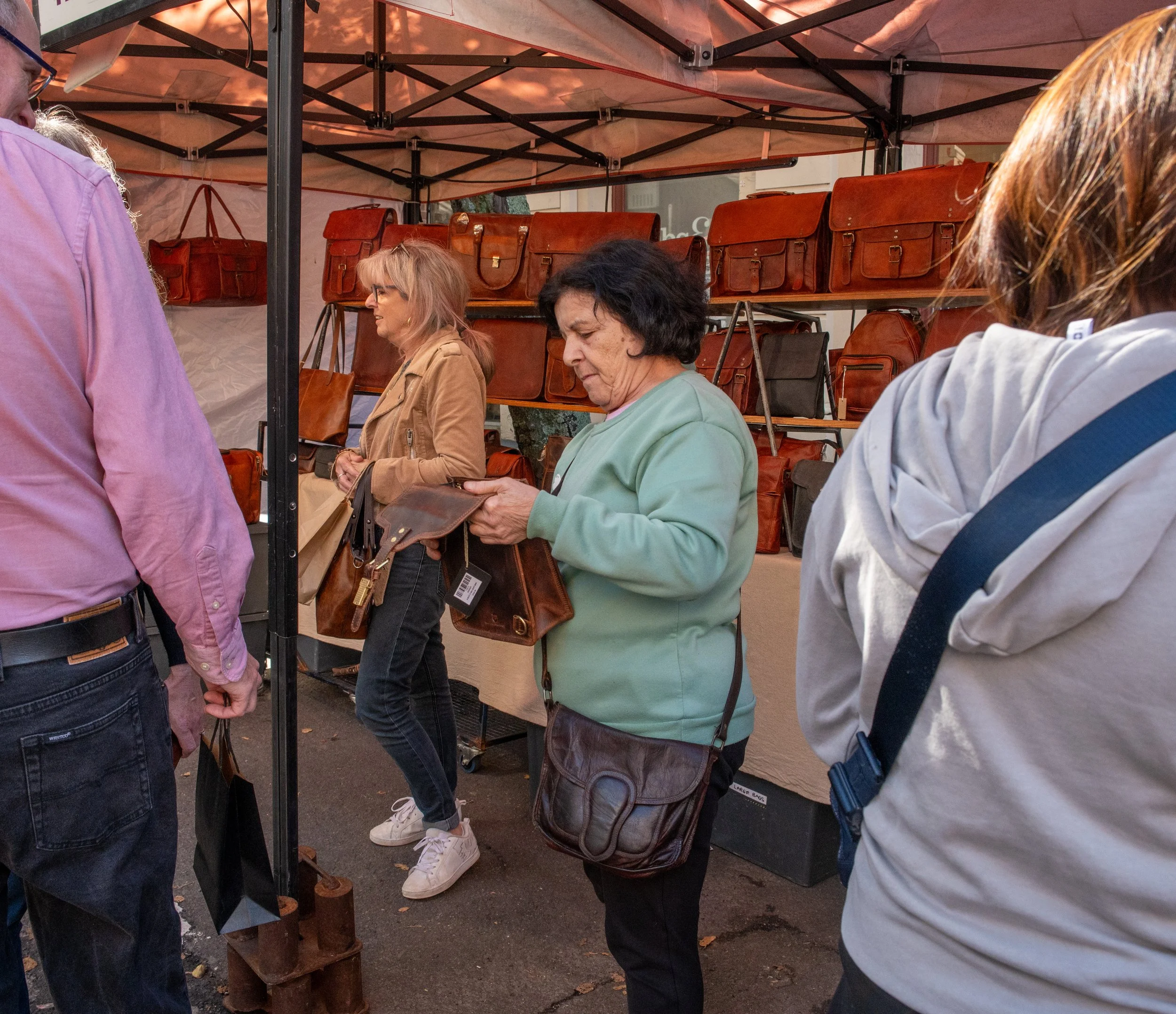 People shopping at an outdoor leather goods stall with handbags on display.