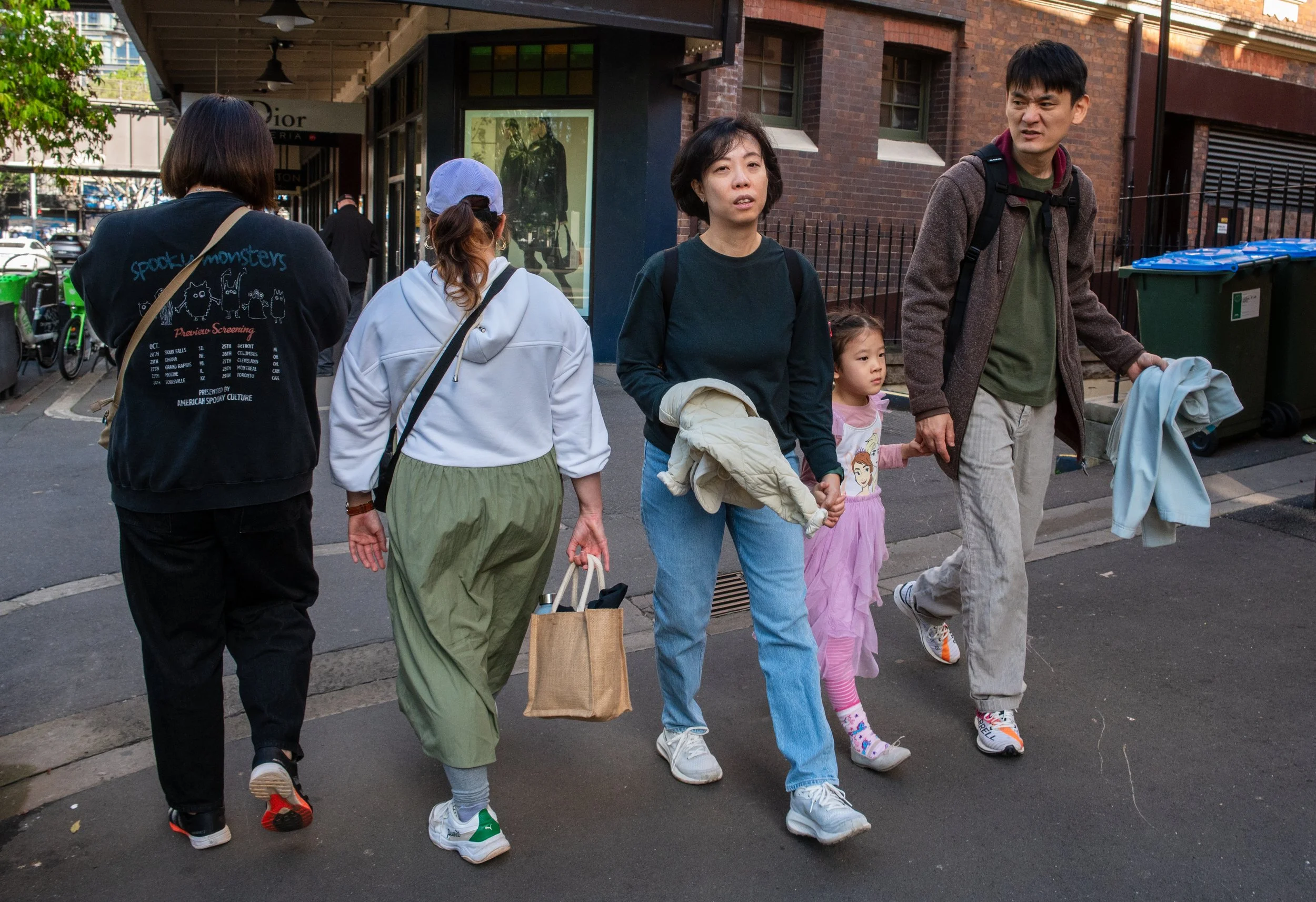 A group of five people, including a young girl, walking on a city sidewalk. The adults are holding shopping bags and their belongings, with buildings and parked bikes in the background.
