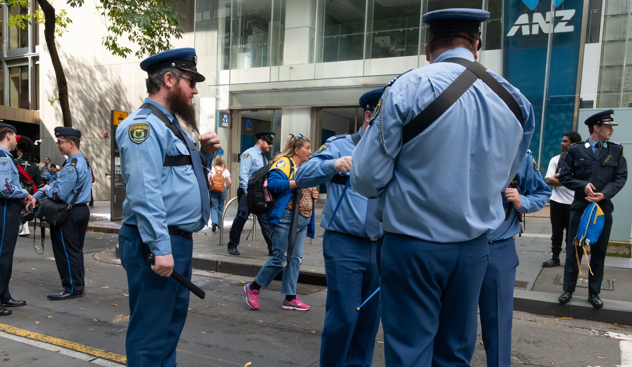 Group of police officers in blue uniforms standing and talking on a city sidewalk, with a few civilians and a modern building in the background.