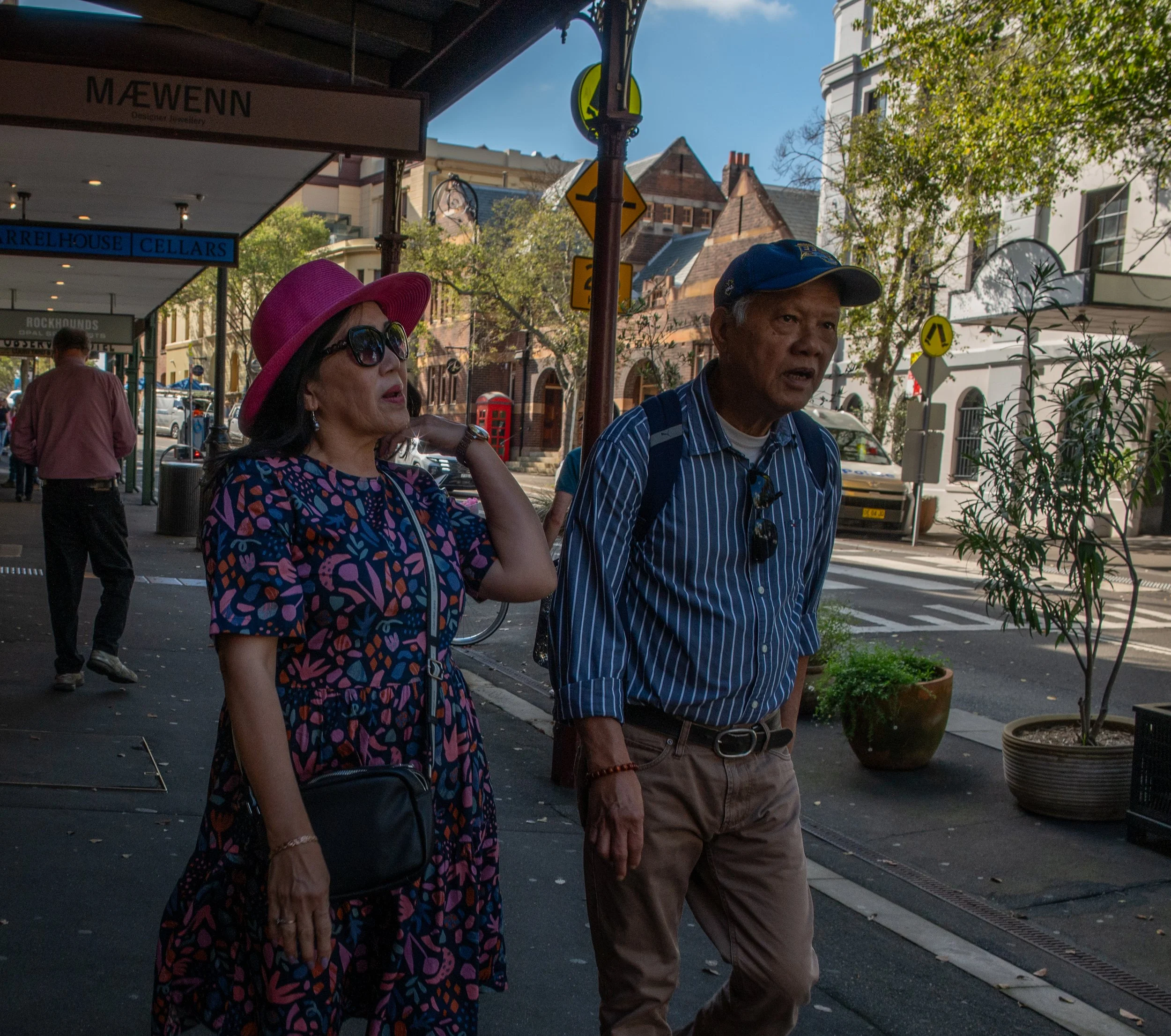 A woman in a purple hat and colorful dress walking alongside a man in a striped shirt and blue cap on a city street with storefronts, trees, and other pedestrians.
