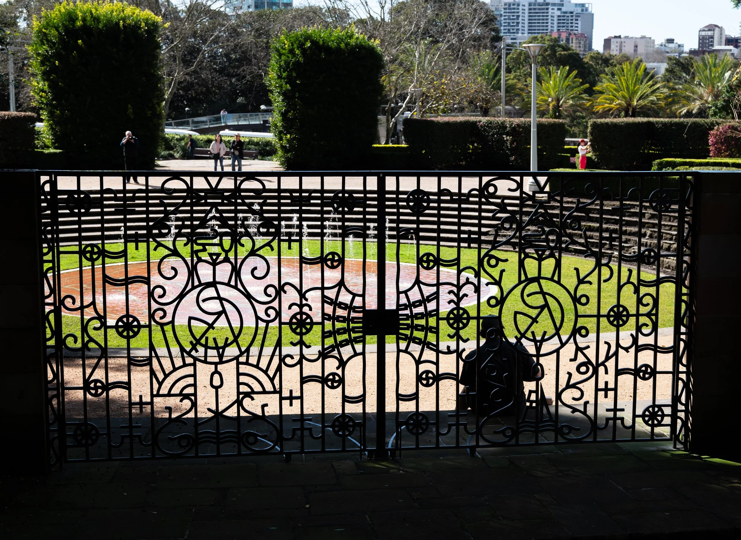 Decorative iron gate leading to a park with steps, lush green bushes, palm trees, and a city skyline in the background.