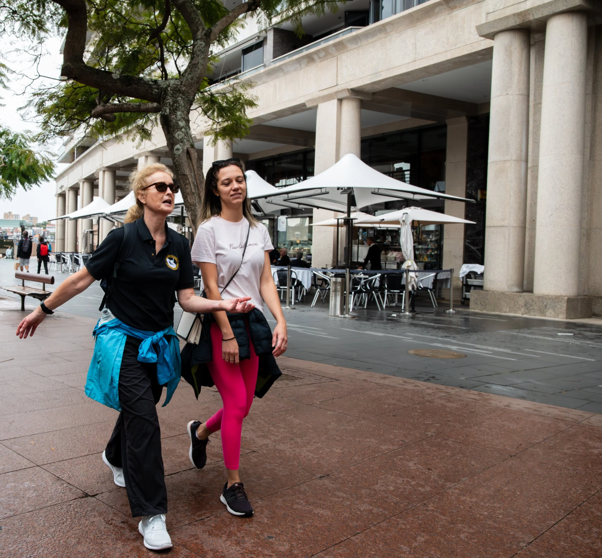 An older woman and a young woman walking outdoors on a city sidewalk near a cafe with outdoor seating, umbrellas, and a large building in the background.