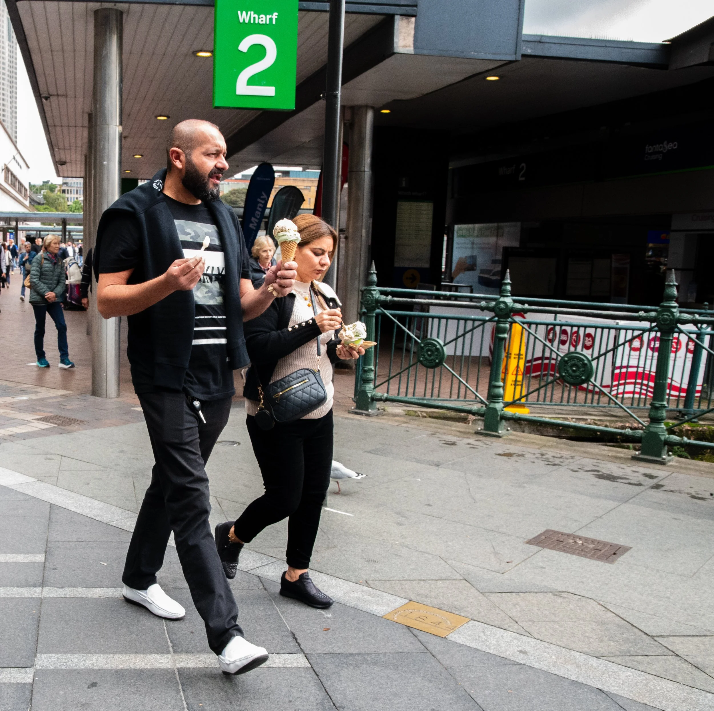 A man and a woman walking on a city sidewalk, each holding ice cream cones, outside a location marked as Wharf 2. There are other pedestrians in the background, and a green sign indicating the location.