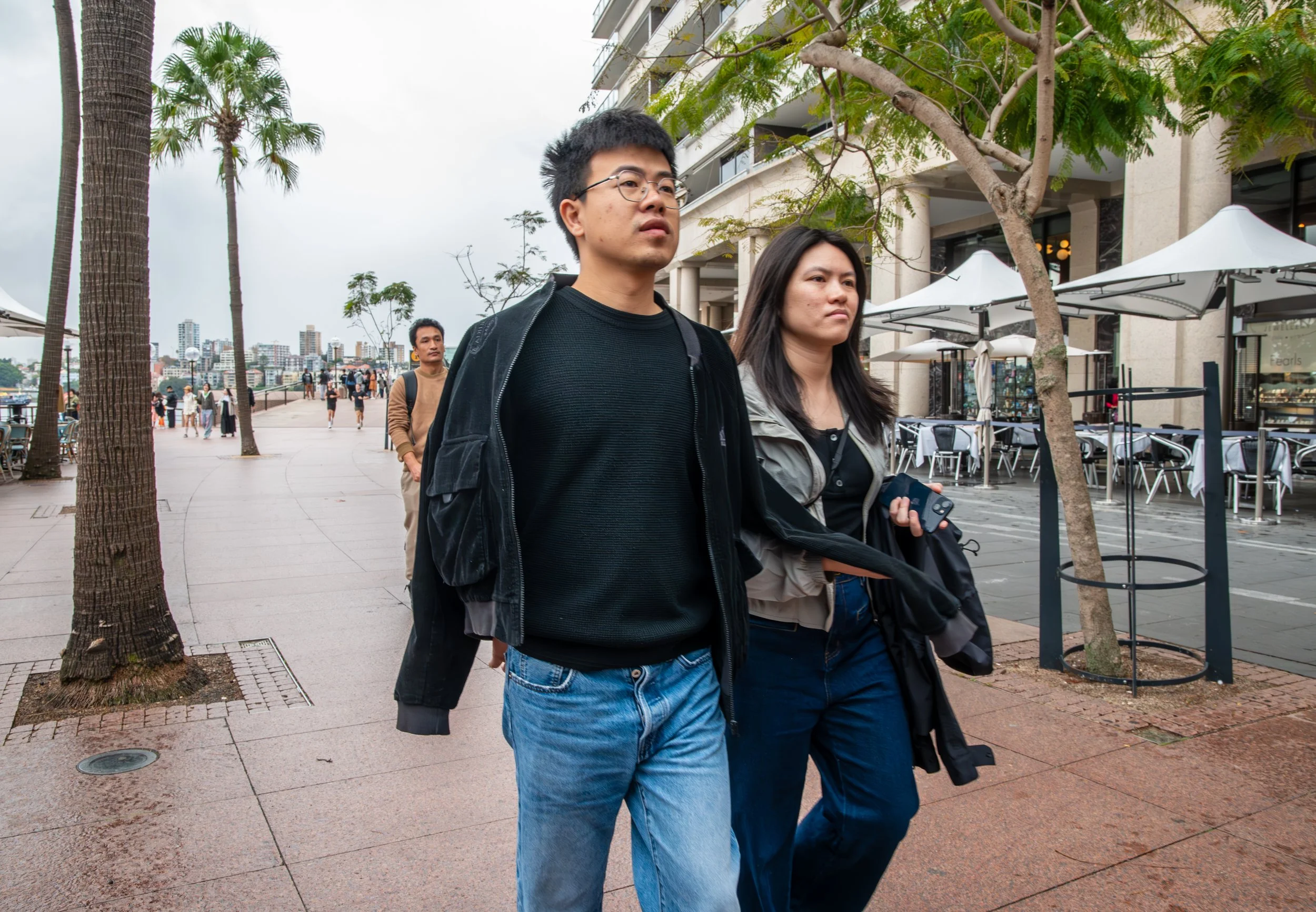 Two young adults walking along a city sidewalk with palm trees and outdoor dining area in the background.