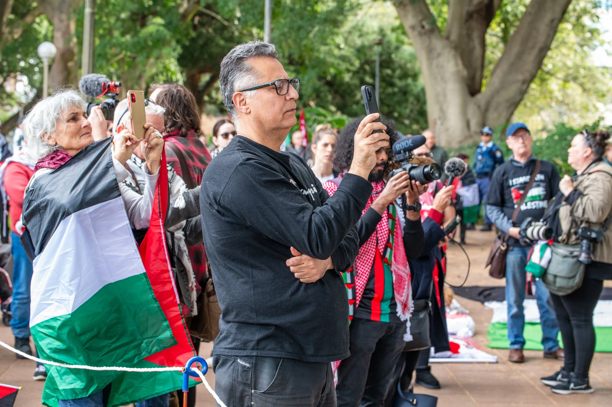 A group of people attending an outdoor event, with some taking photos or videos with their phones and cameras, surrounded by trees and greenery.