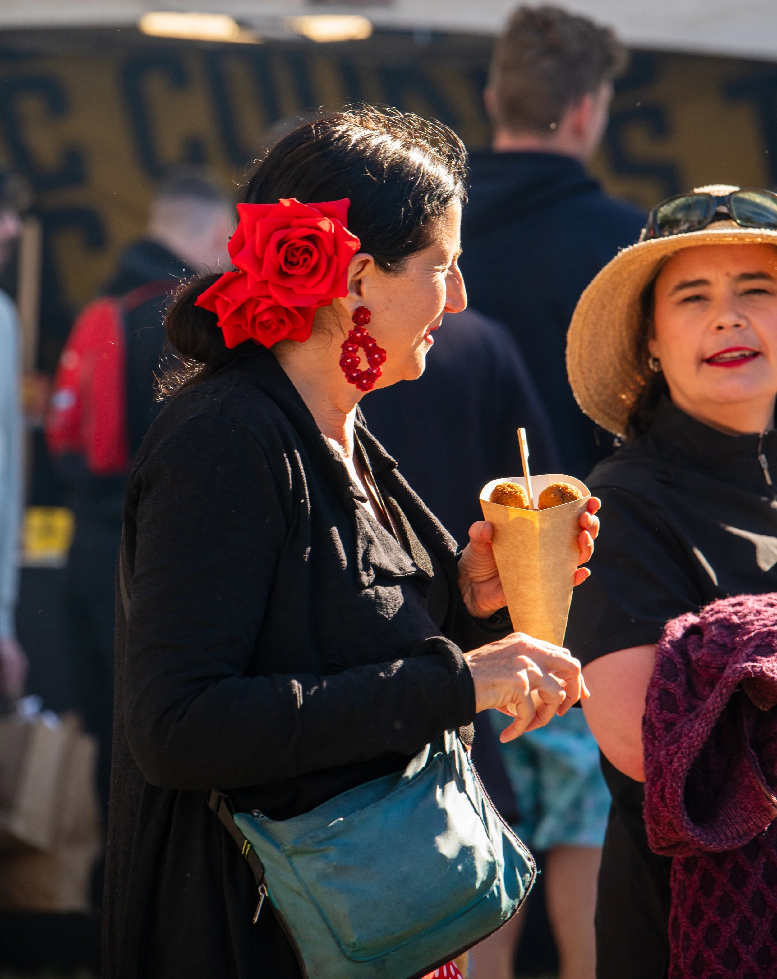 A woman with dark hair, wearing large red rose hair accessories and red earrings, is holding a paper cone filled with food at an outdoor event. She wears a black outfit and has a teal purse across her shoulder. She is talking to another woman wearing a wide-brimmed straw hat and black top.
