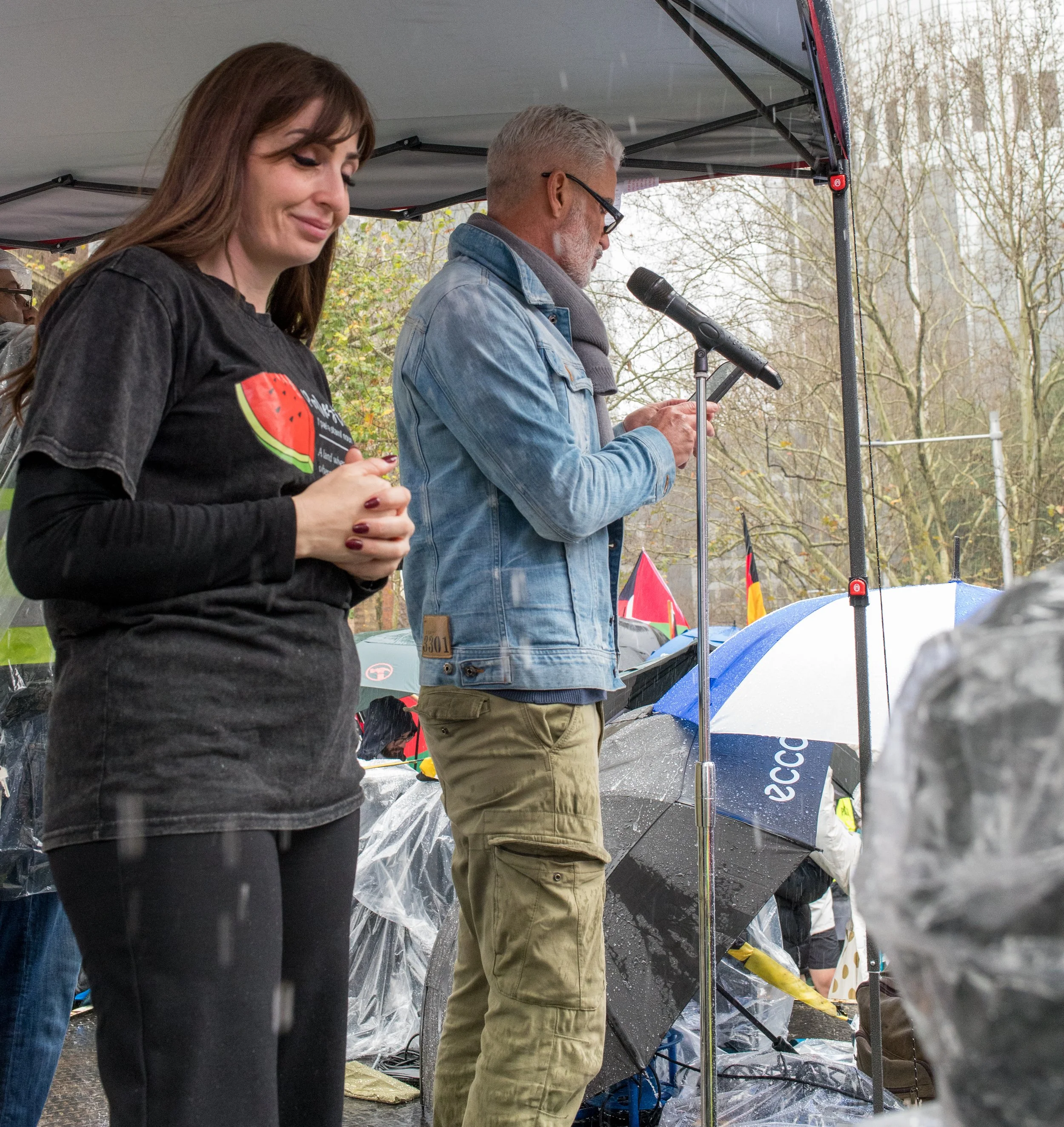 A woman with long brown hair, wearing a black t-shirt with a watermelon graphic, holding her hands together, and a man with gray hair and glasses, wearing a denim jacket, speaking into a microphone at an outdoor event during rain, with umbrellas and trees in the background.