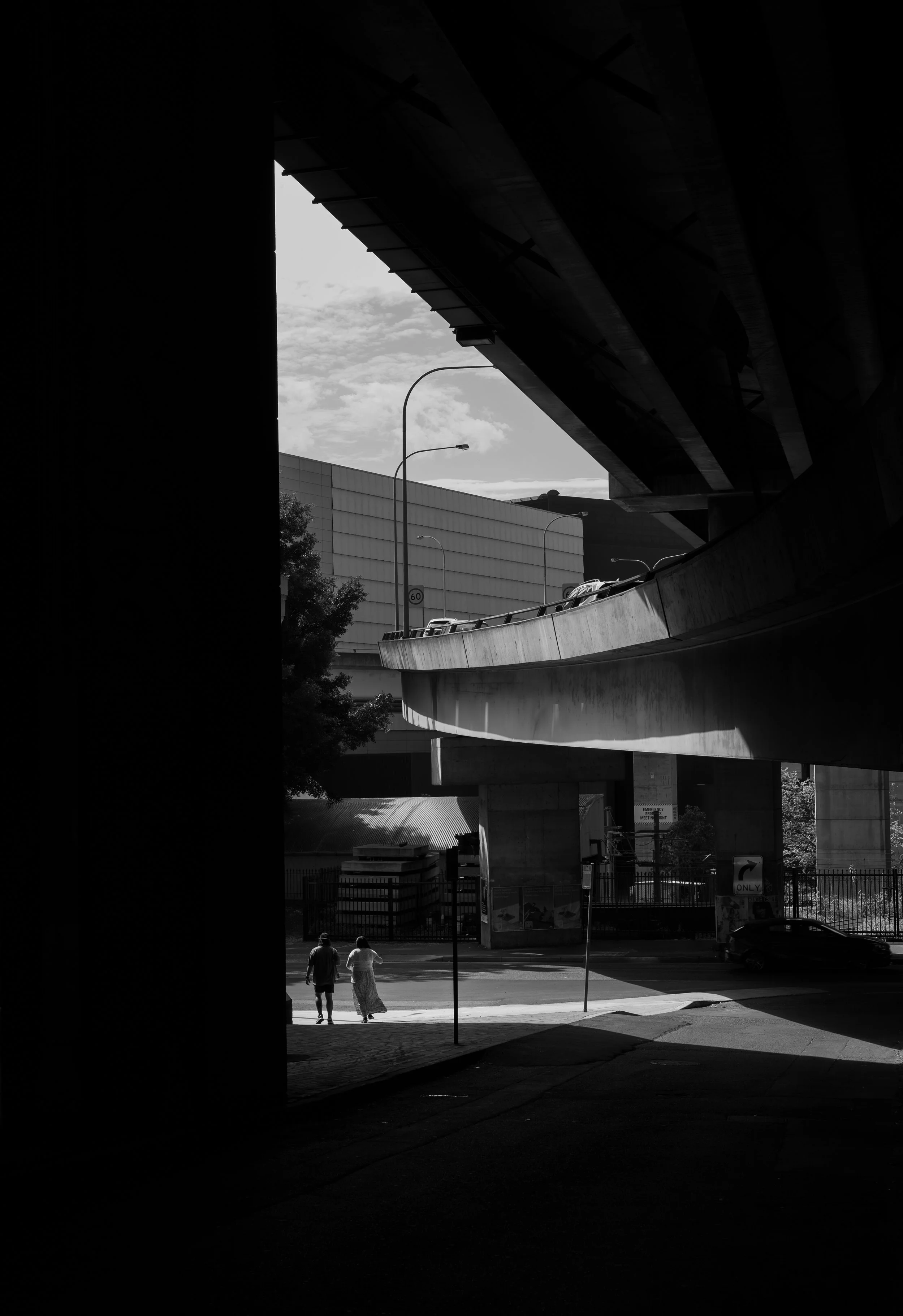 A black and white photo of a city street scene taken from a shadowed area, showing two people crossing the street beneath an overpass with cars on it, and buildings in the background.