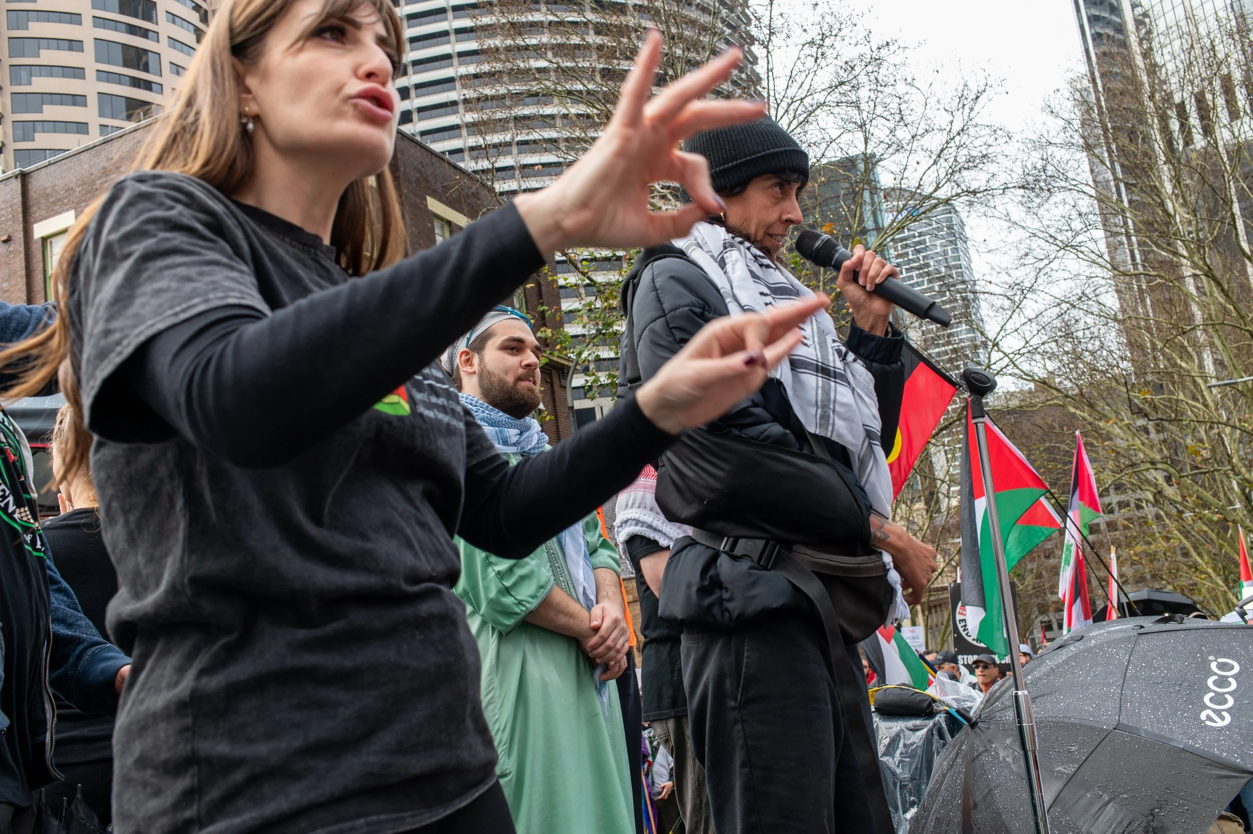 People protesting outdoors, some speaking into a microphone, with flags in the background, cloudy sky, and trees.