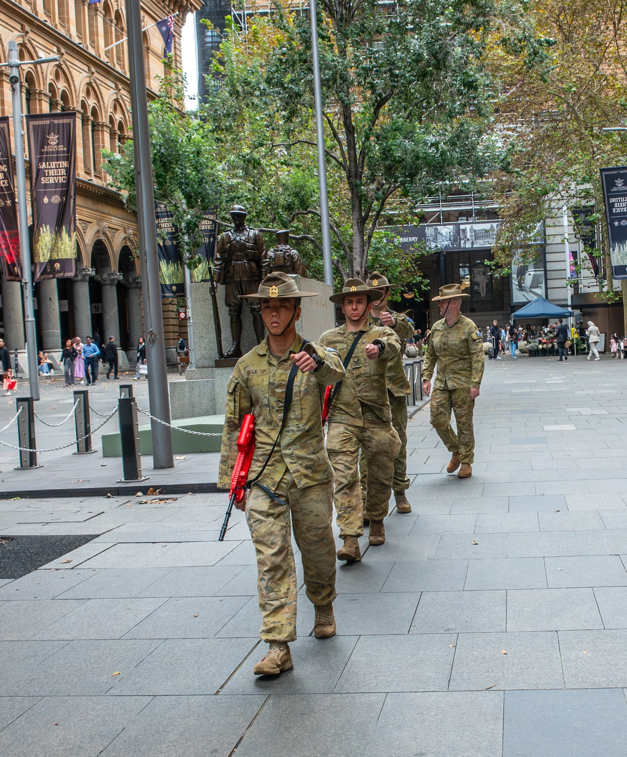 Four soldiers in camouflage uniforms walking down a city sidewalk with historic buildings and trees in the background.