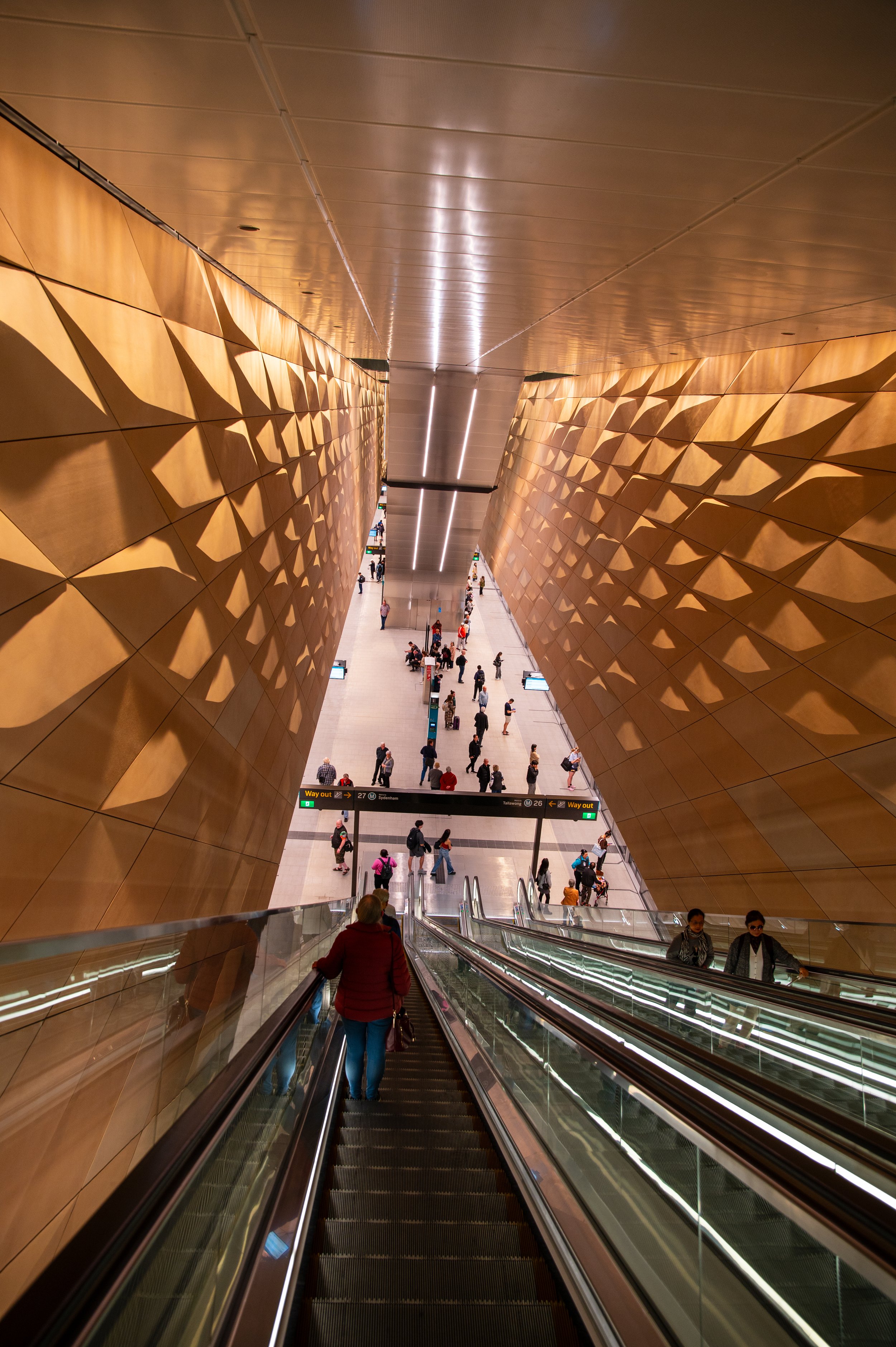 A view of an escalator descending inside a modern airport terminal with gold-colored walls featuring geometric patterns and people waiting in line below.