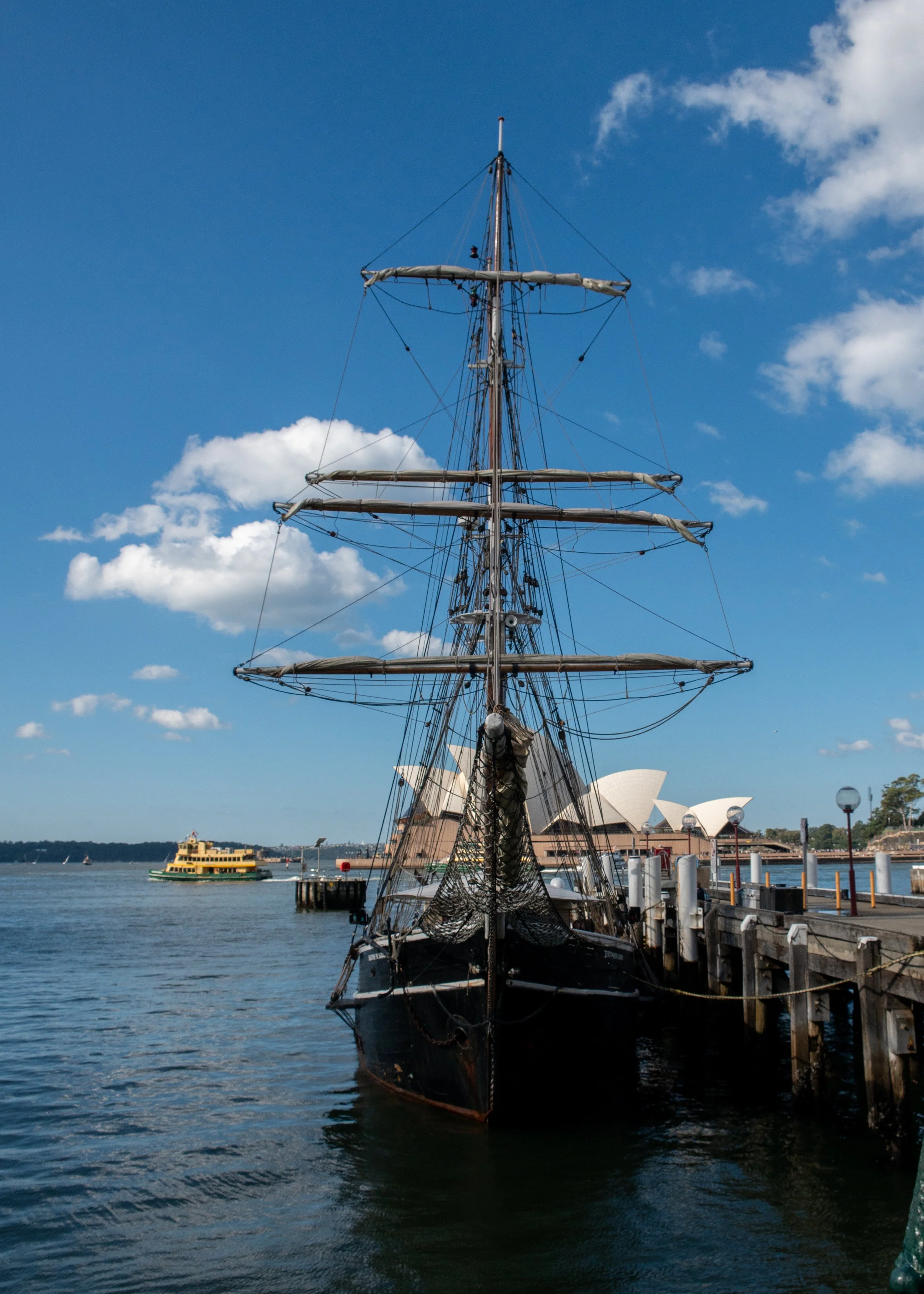 Old black sailing ship docked at a harbor, with the Sydney Opera House visible in the background under a partly cloudy sky.