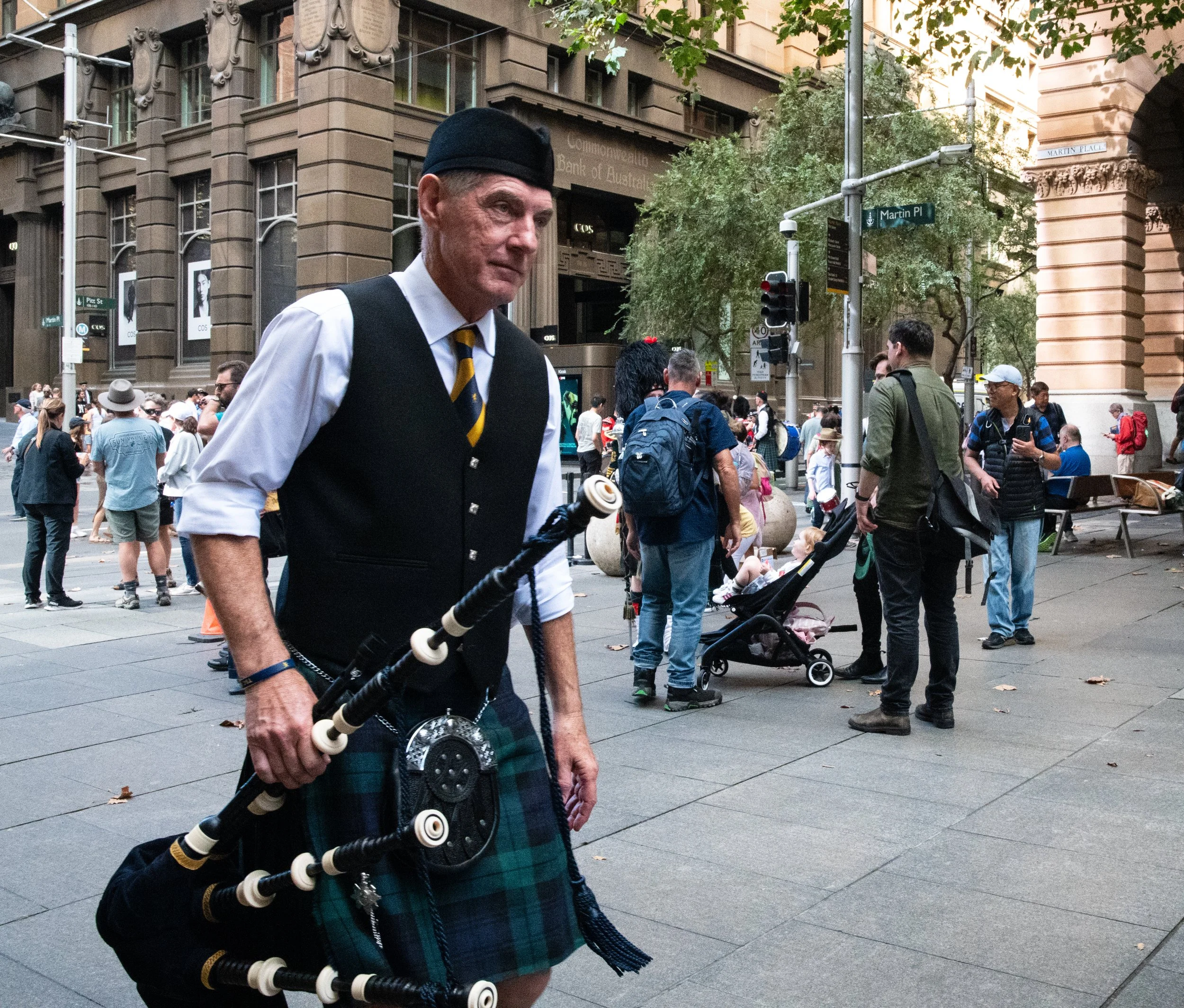 A man in traditional Scottish attire playing bagpipes on a busy city street. The background shows people walking, some with children and strollers, and tall buildings with multiple windows and architectural details.