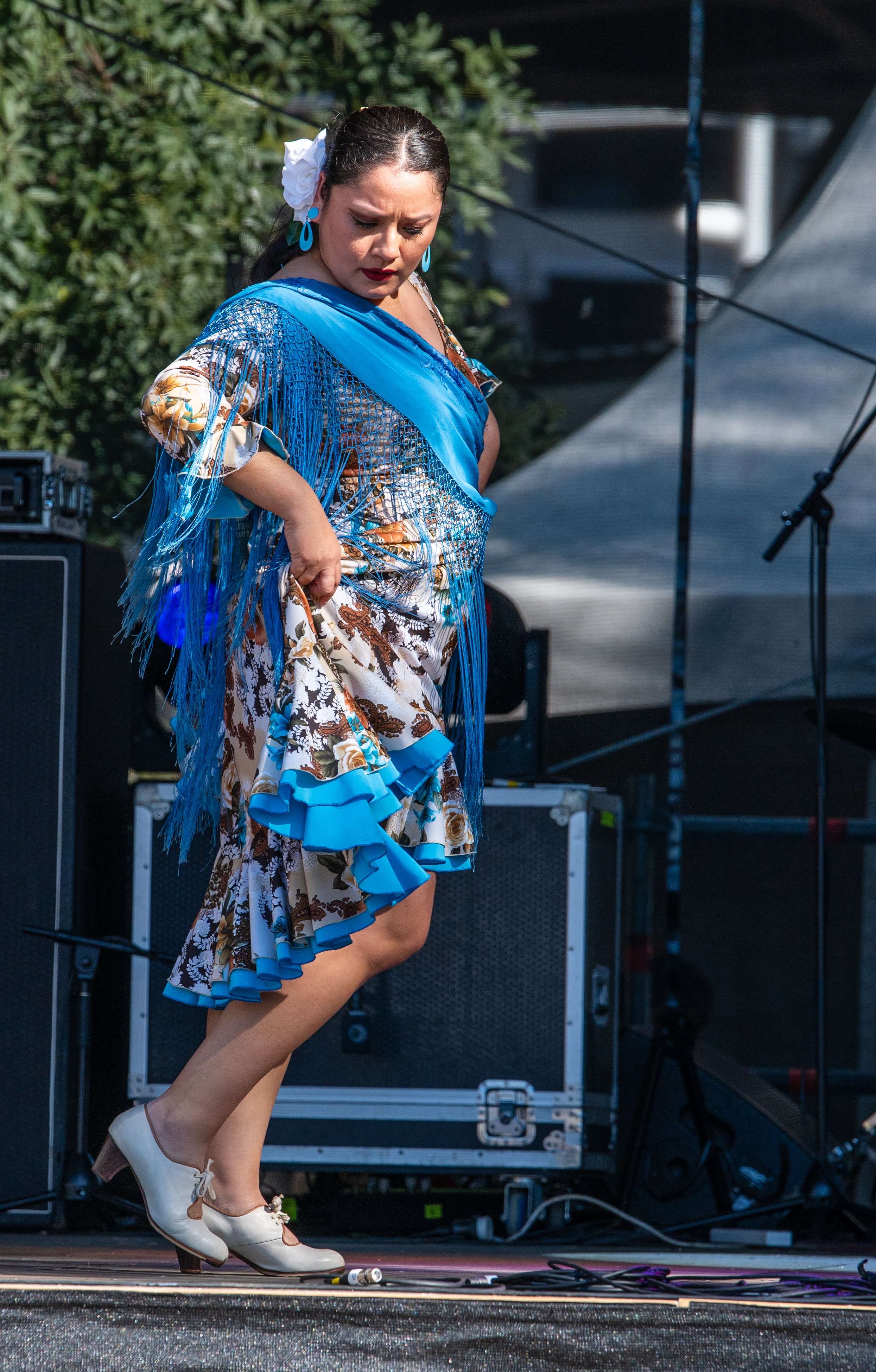 A woman performing a traditional Mexican dance on stage, dressed in a vibrant floral dress with ruffles and blue fringe, wearing beige dance shoes, and holding her skirt while looking down.