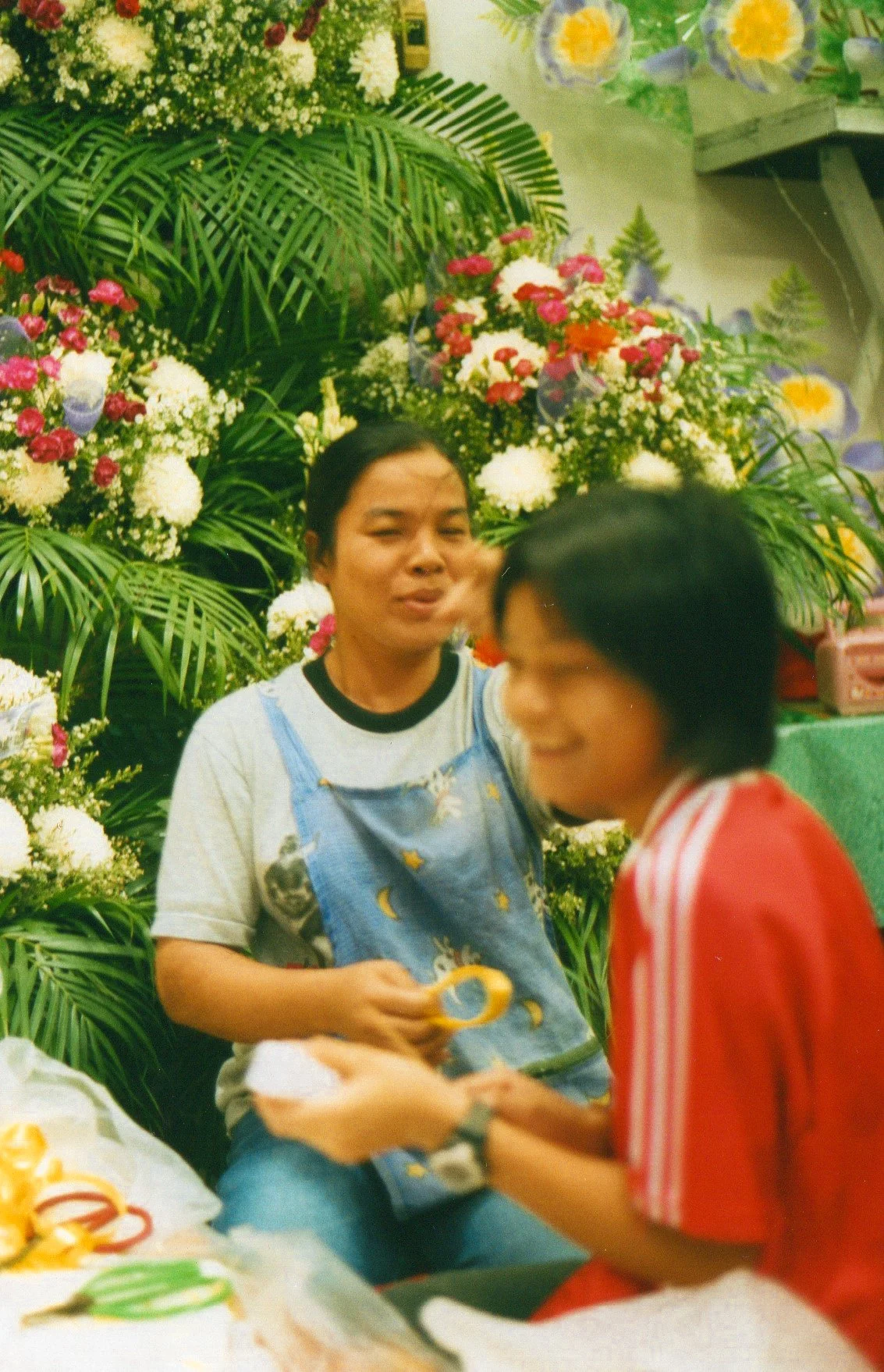 Two women sitting in a flower shop, surrounded by colorful floral arrangements and greenery, engaging in conversation.