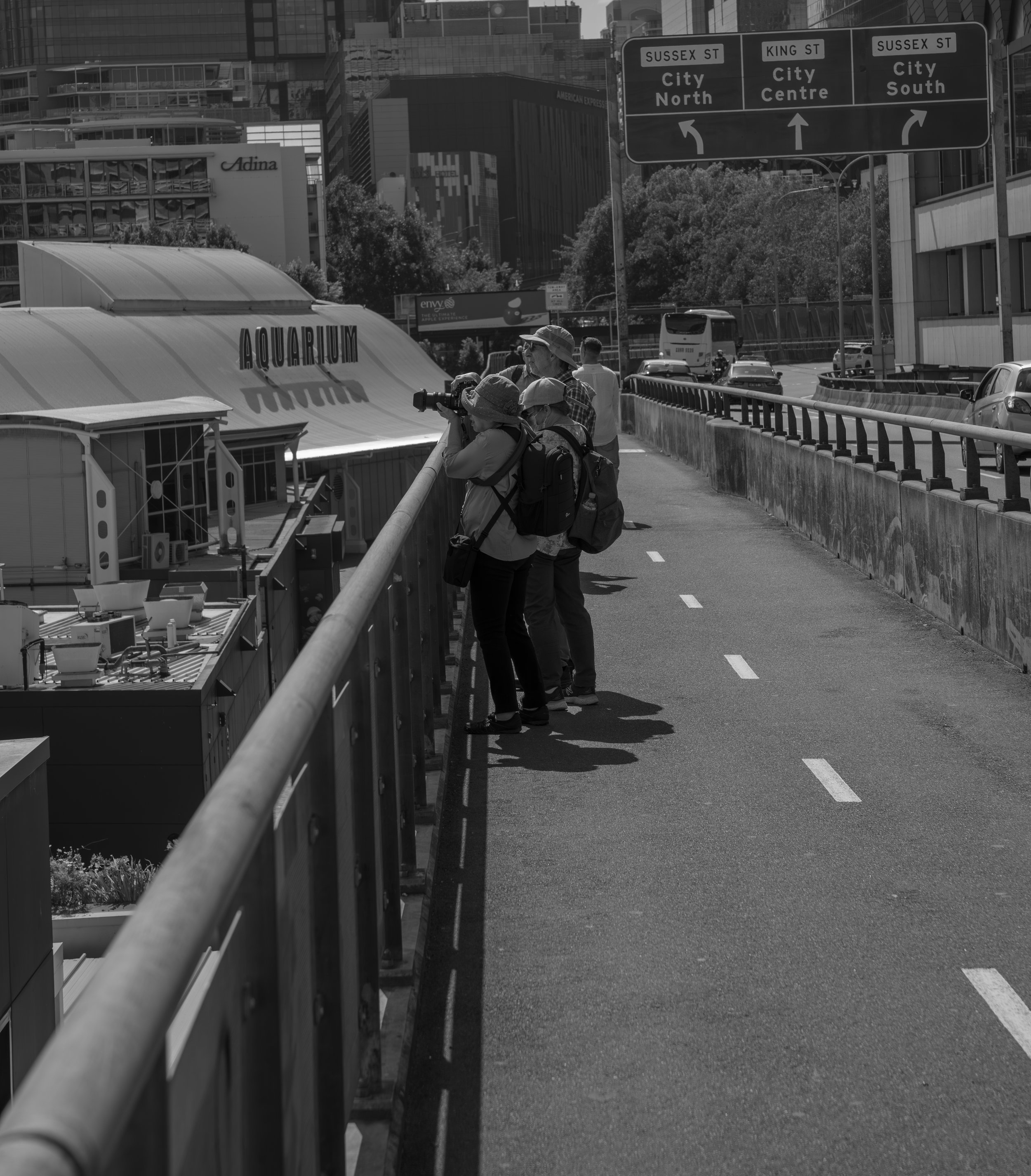 Four people observing through binoculars from a pedestrian bridge in an urban area, with a sign overhead indicating directions to Sussex Street, King Street, and City South.