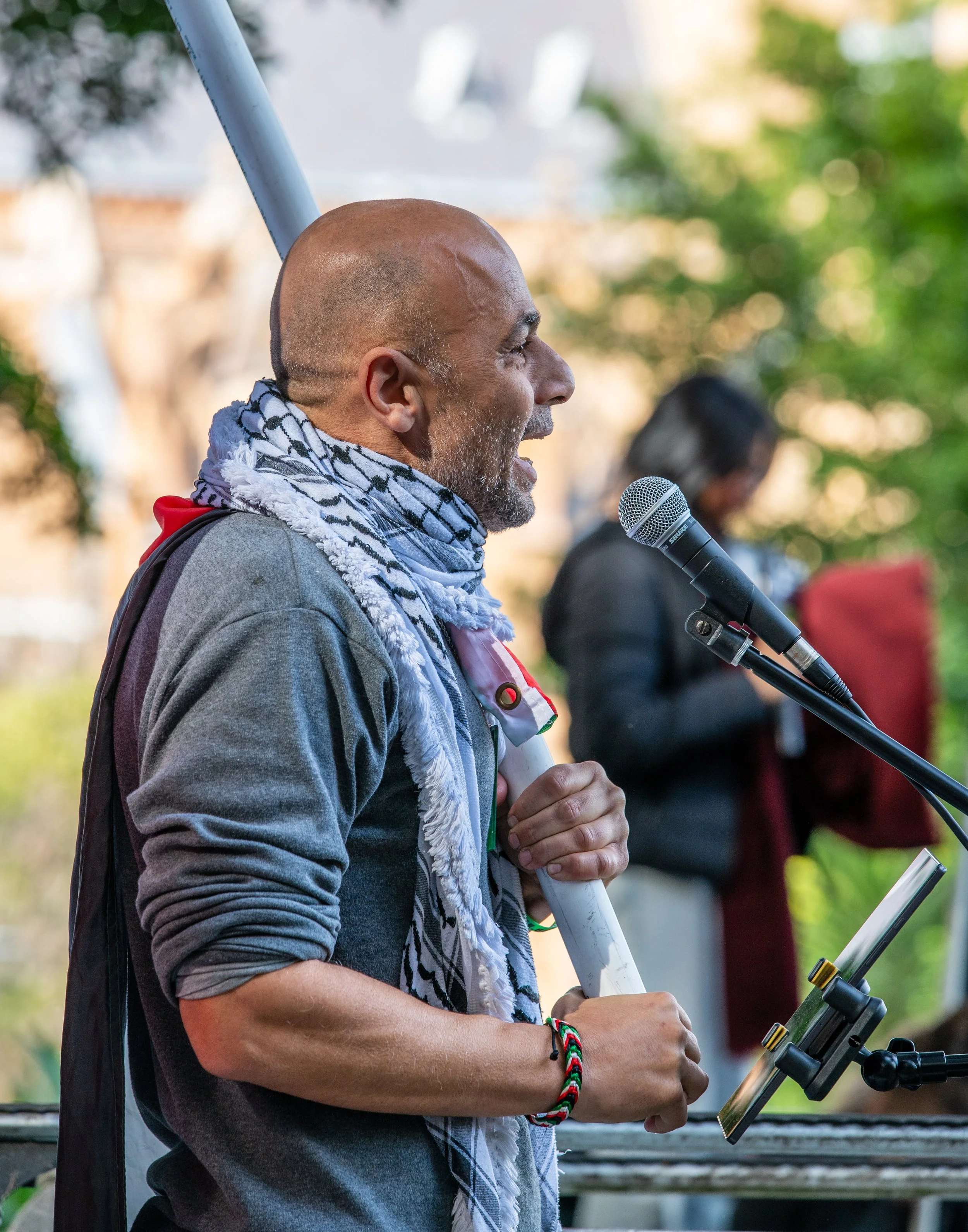A man speaking into a microphone at an outdoor event, holding a flagpole with a scarf around his neck, with a woman in the background reading from a book or paper.